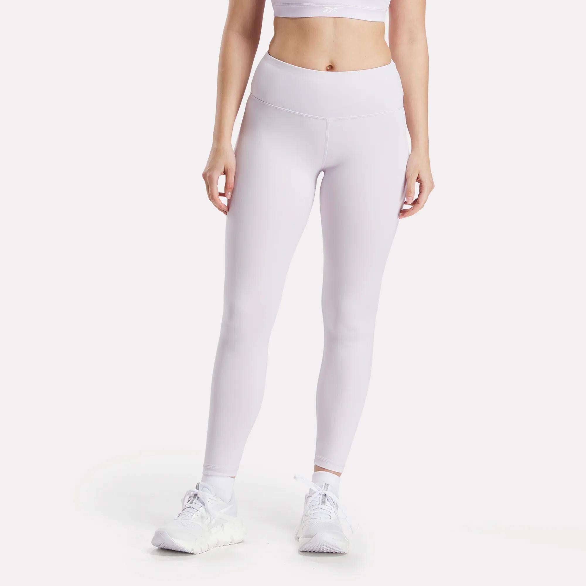A person wearing a white sports bra, Reebok Women's Core High-Rise Leggings, and white athletic shoes stands against a plain white background with their arms relaxed at their sides.