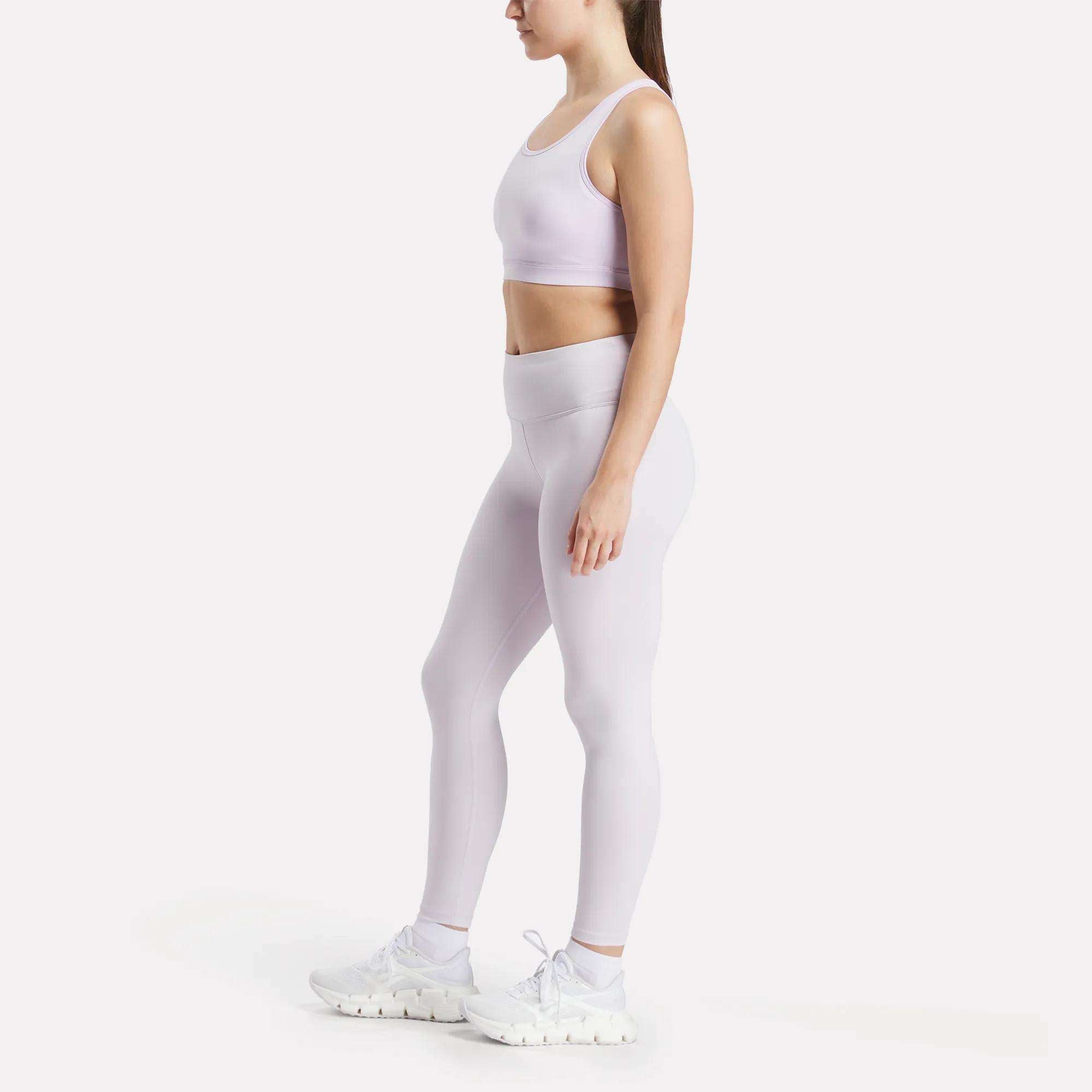 A woman stands in profile against a white background, wearing Reebok Women's Core High-Rise Leggings in light purple with a matching sports bra and white athletic shoes, featuring RBK-DRY technology.
