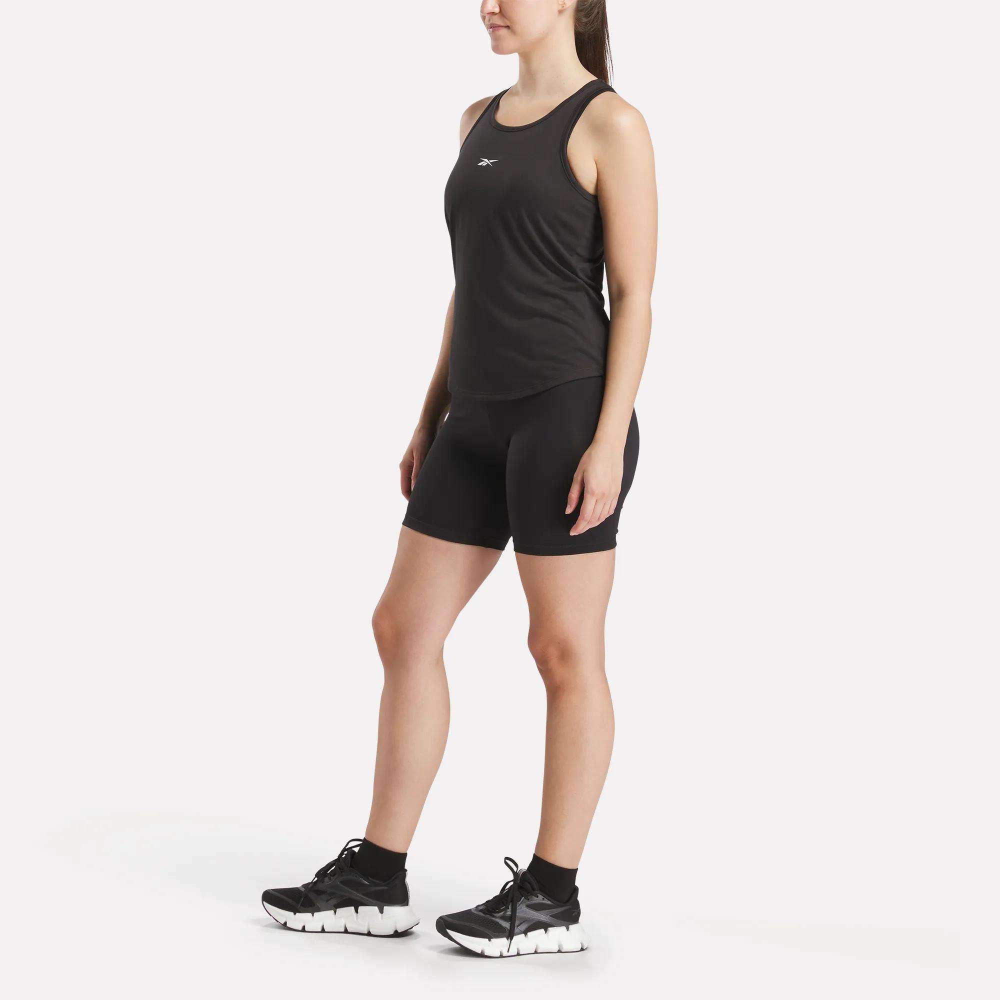 A woman stands against a white background wearing a black Reebok Women's Core Tank Top, black bike shorts, black socks, and black women's Reebok athletic shoes with thick white soles.