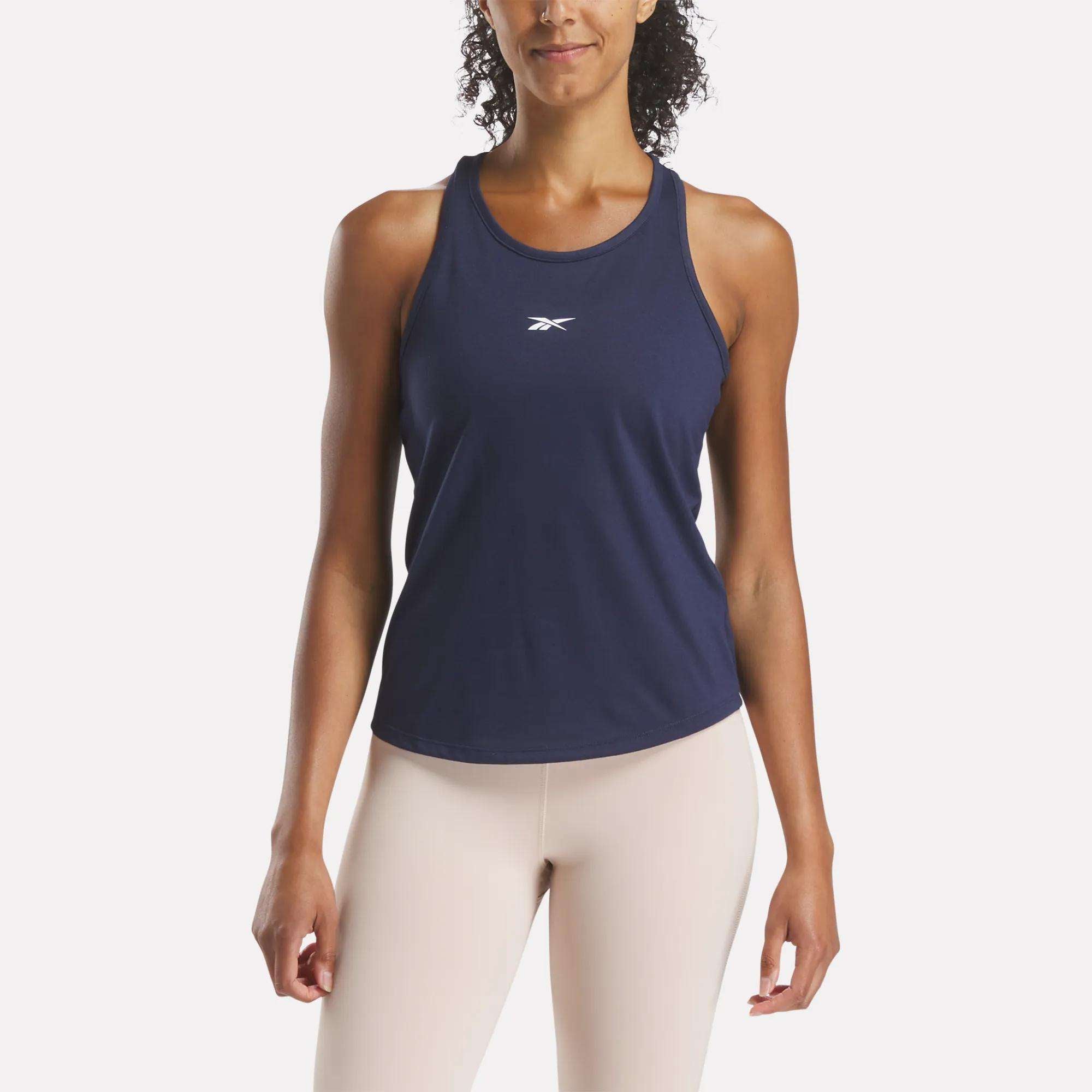 A woman in a slim fit, navy blue Reebok Women's Core Tank Top with sweat-wicking fabric and a small white logo on the chest pairs it with light beige leggings, standing against a plain white background.