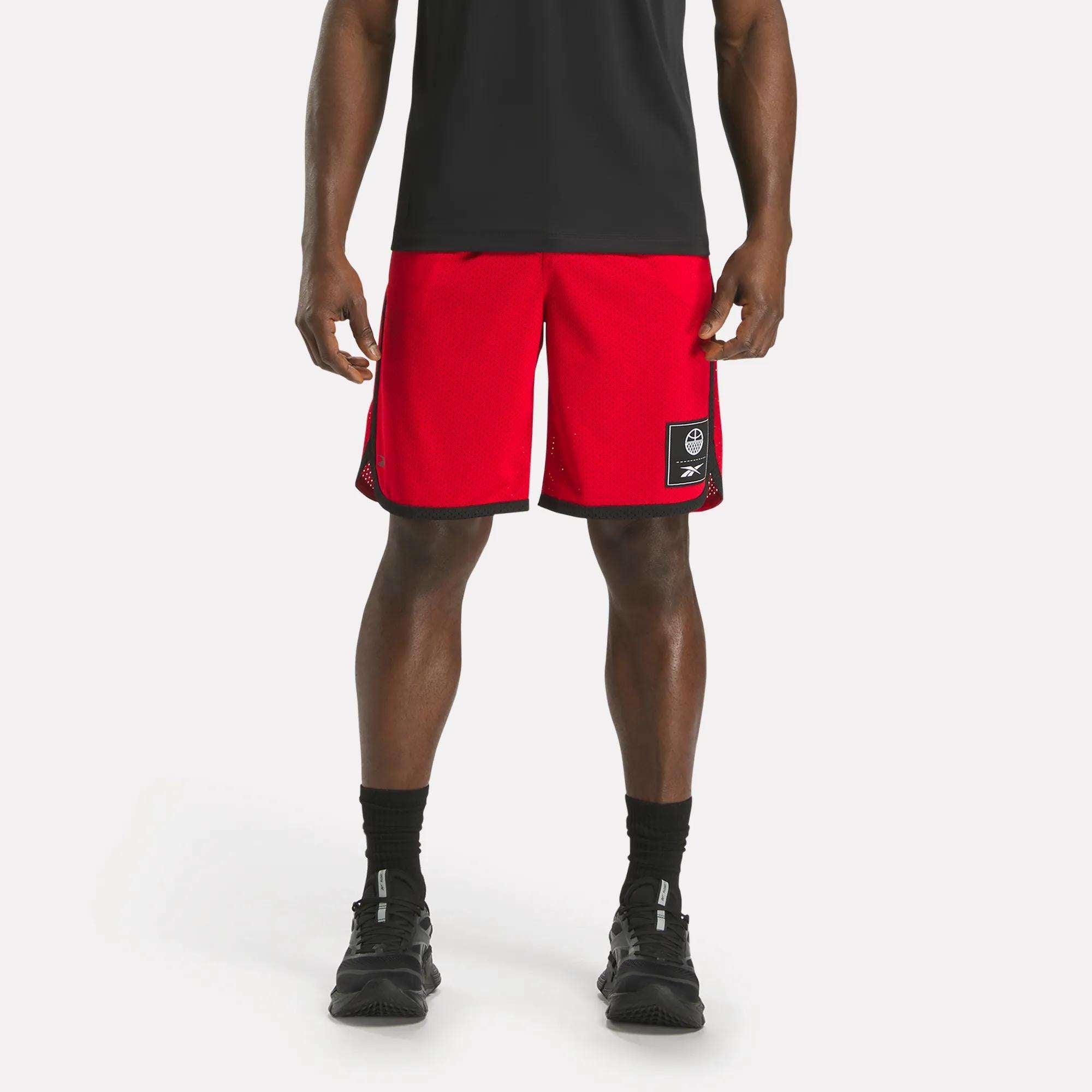 A person wearing a black shirt, Reebok Men's Basketball Mesh Shorts in red with RBK-DRY tech and a patch on the left leg, black socks, and black sneakers stands against a plain white background.