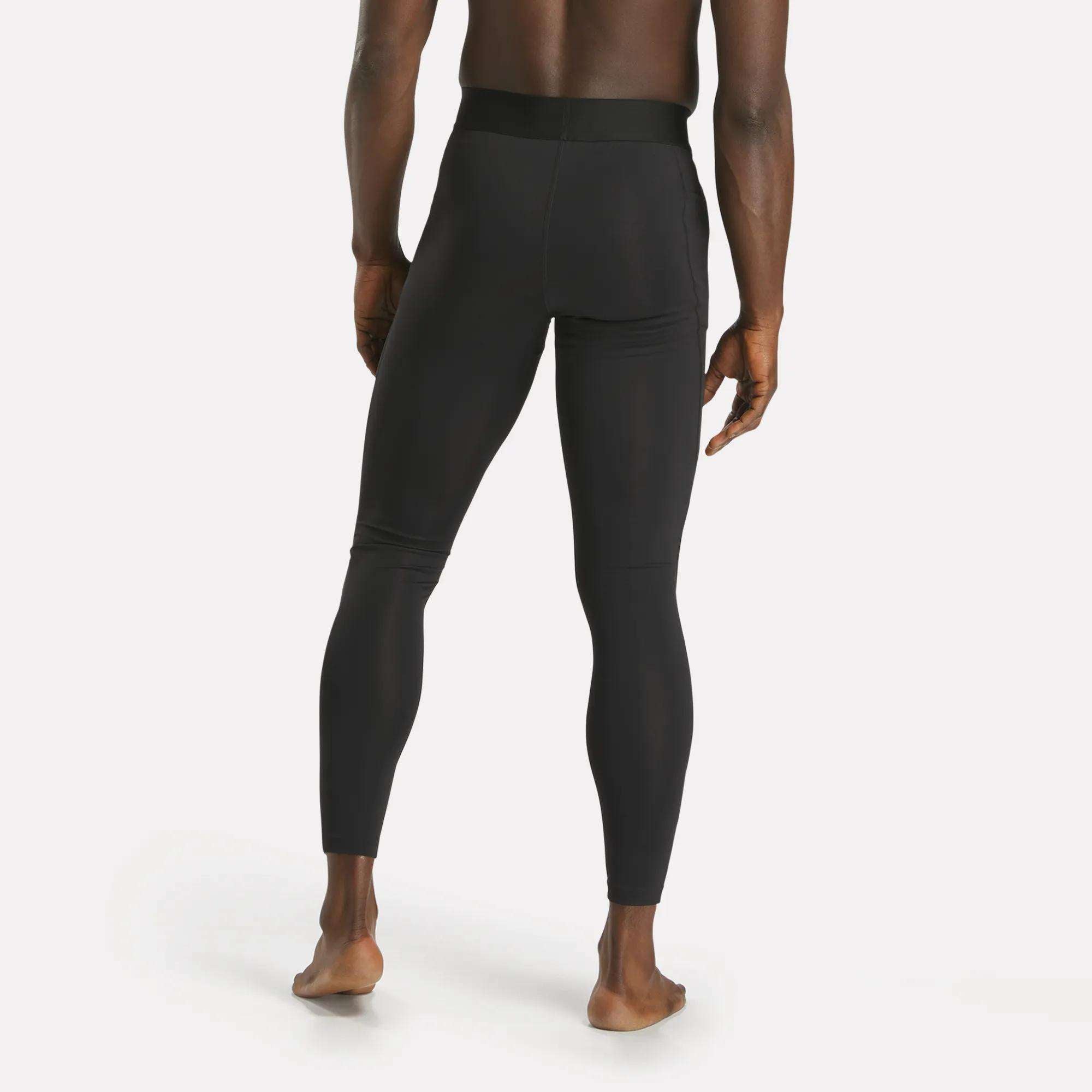 A barefoot person seen from behind wears Reebok Men's Core Training Tights in black, standing on a white background.