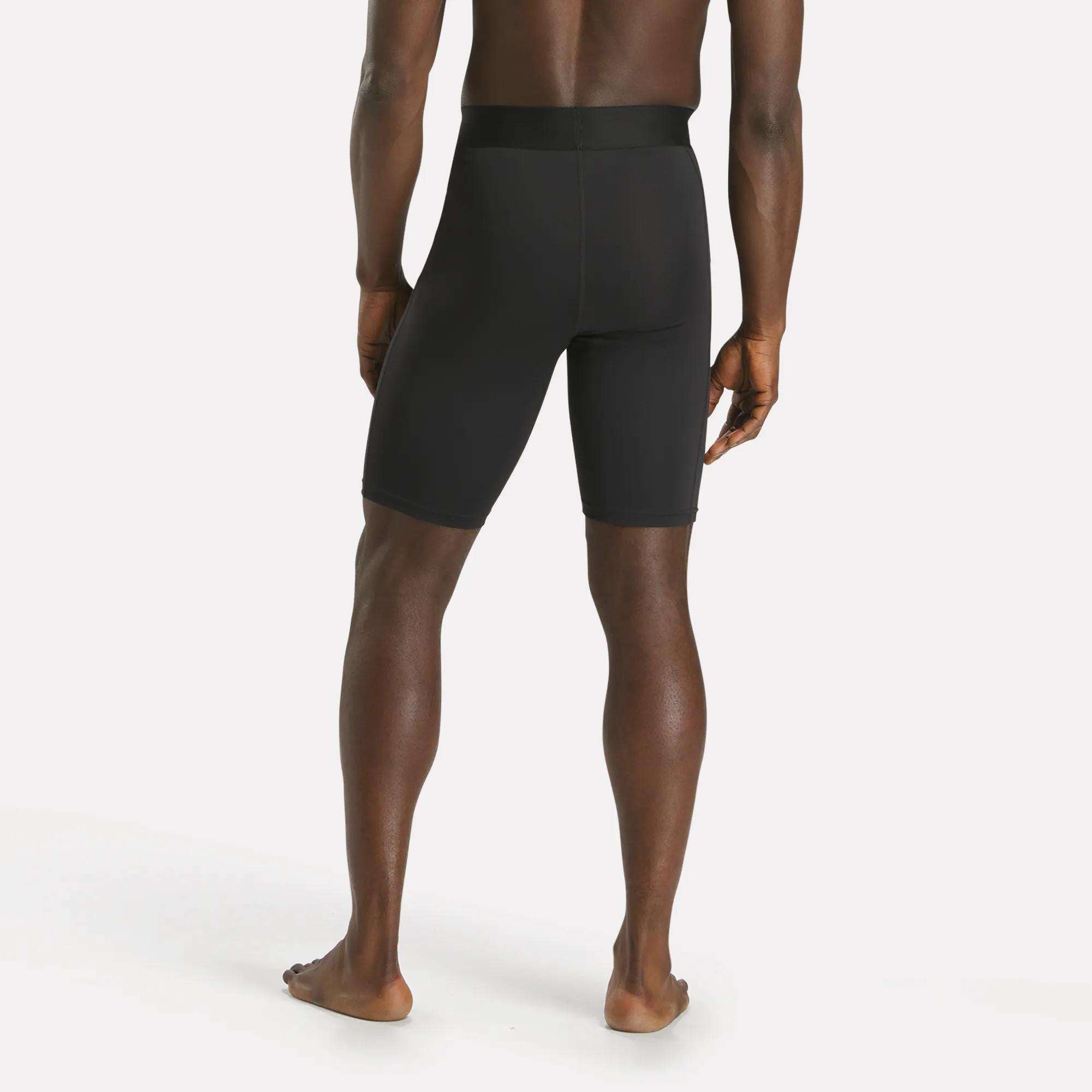 A man stands barefoot, facing away with arms at his sides and legs slightly apart, modeling Reebok Men's Training Short Tights made from recycled polyester against a plain white background.