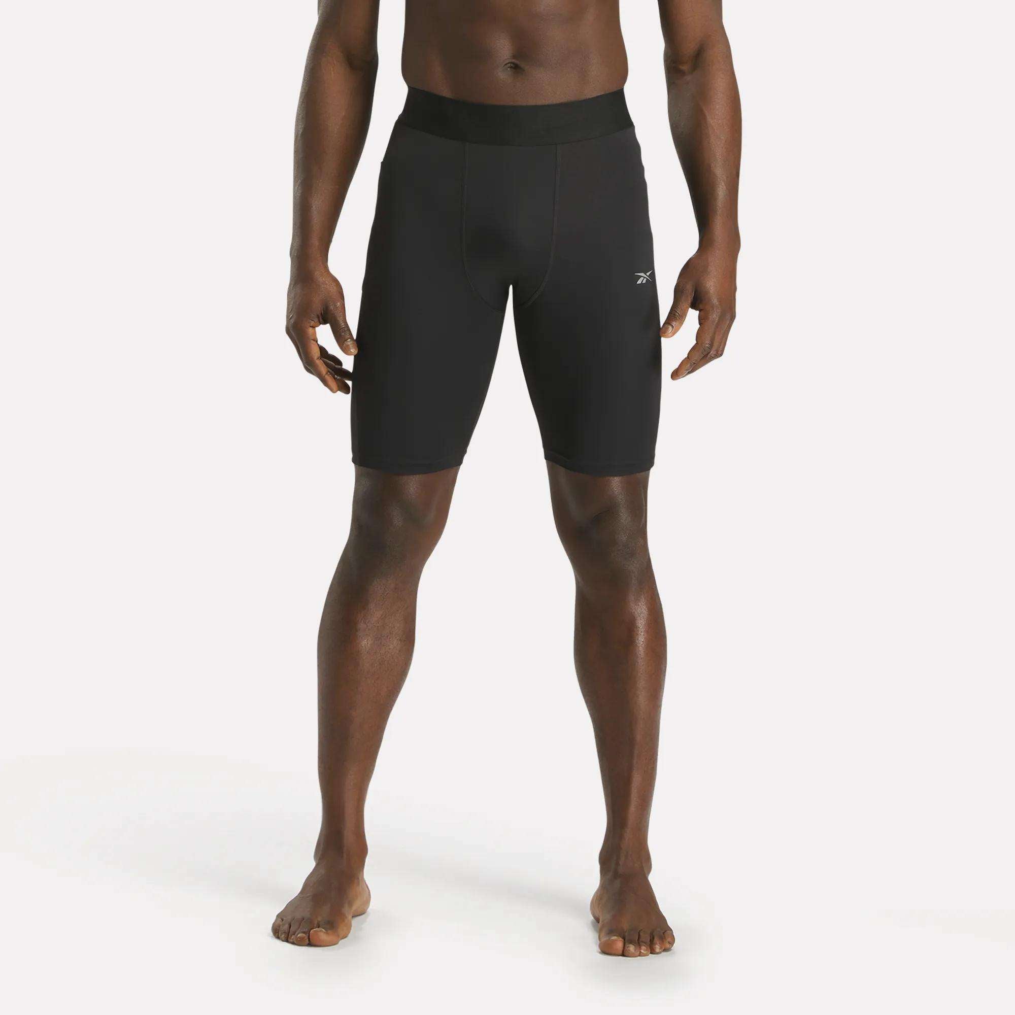 A barefoot man stands facing forward in Reebok Men's Training Short Tights, featuring a black fitted design made from recycled polyester, set against a plain white background.