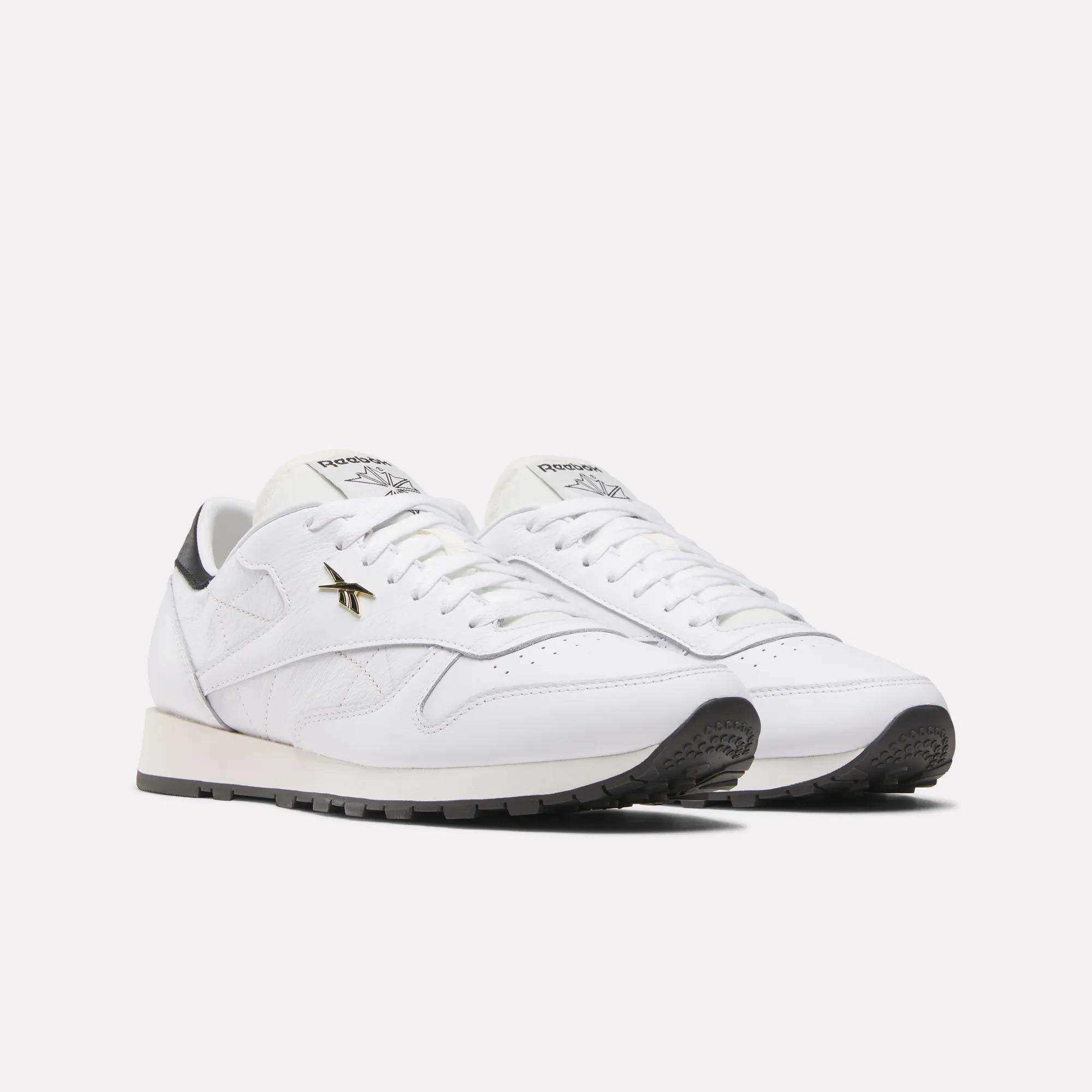 The Reebok Classic Leather Shoes showcase a retro running style with a white leather upper, black soles, gold logo accents, and white laces, set against a plain light background.