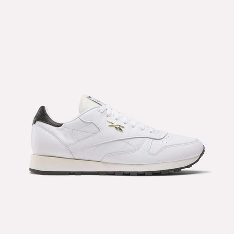 The Reebok Classic Leather Shoes feature a white leather upper, black heel detail, gold Reebok logo on the side, white laces, and a black sole, shown against a light background.
