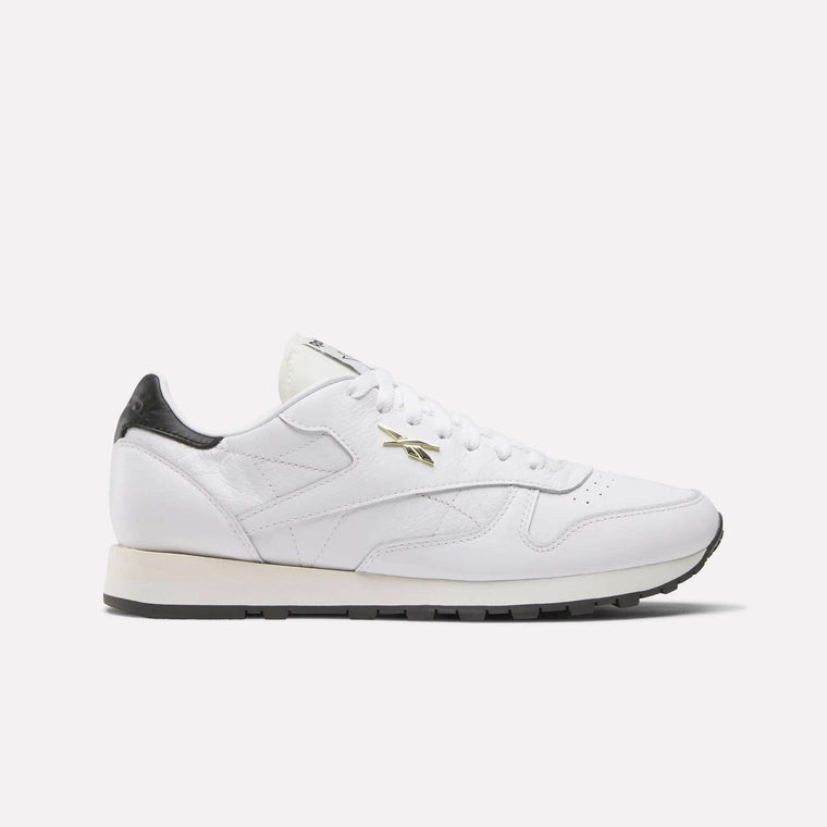 The Reebok Classic Leather Shoes feature a white leather upper, black heel detail, gold Reebok logo on the side, white laces, and a black sole, shown against a light background.