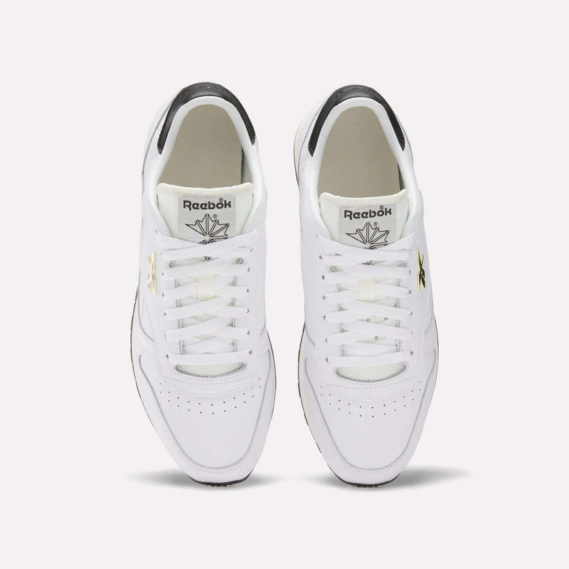 Top view of Reebok Classic Leather Shoes featuring a leather upper, white laces, black and gold logo accents, and a grey heel detail, set against a plain light background.