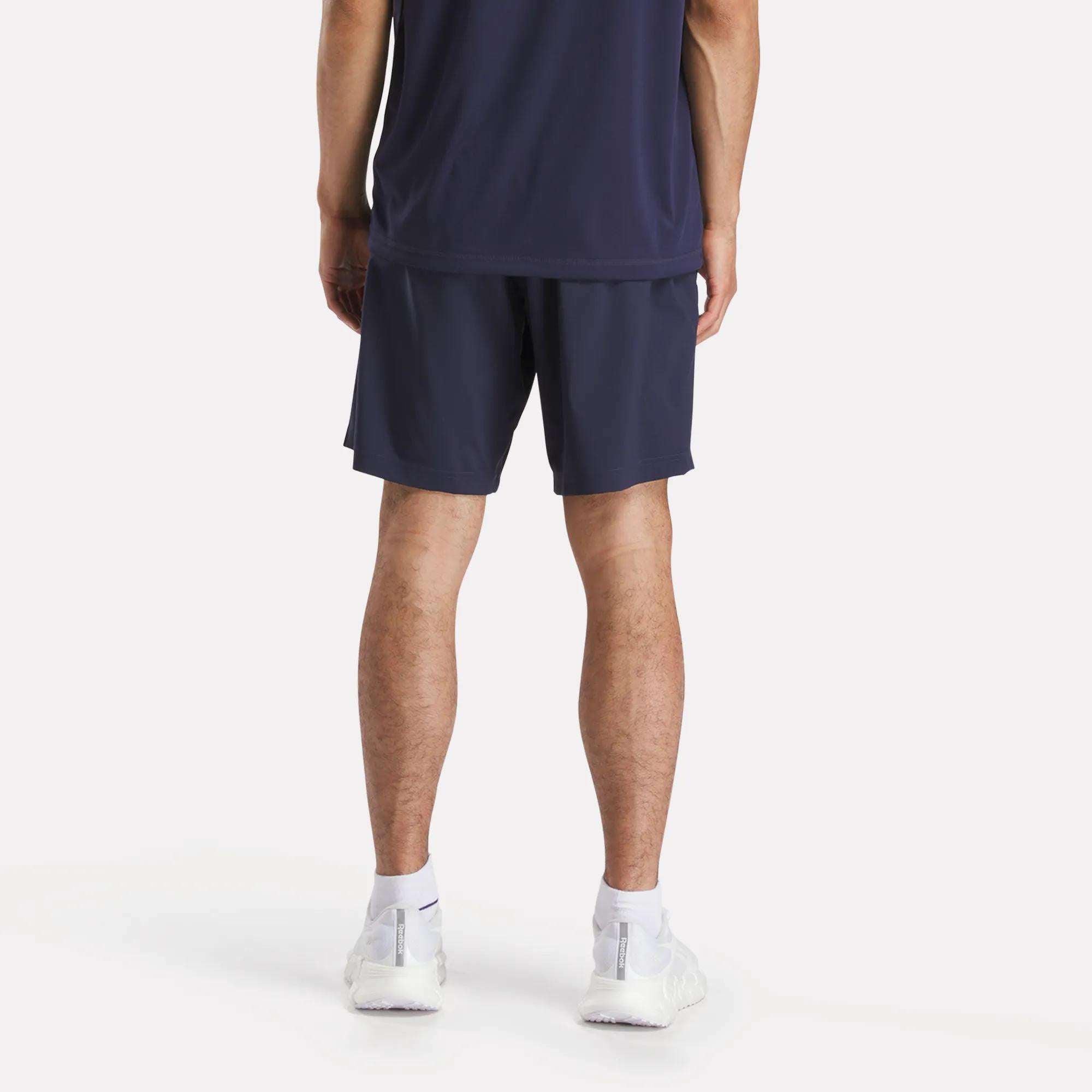A person stands facing away from the camera, wearing a navy athletic shirt and Reebok Men's Workout Ready Woven Shorts, paired with white socks and sneakers against a plain white background.