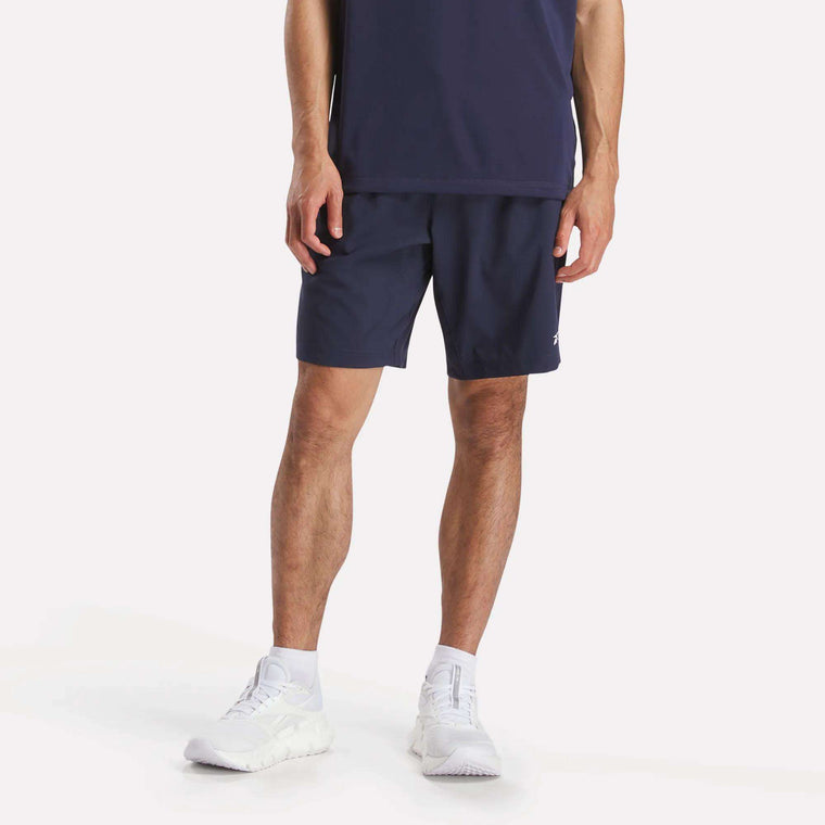 A person stands against a plain background, wearing a navy blue shirt, white athletic shoes, white socks, and Reebok Men's Workout Ready Woven Shorts. The image is cropped at the shoulders.