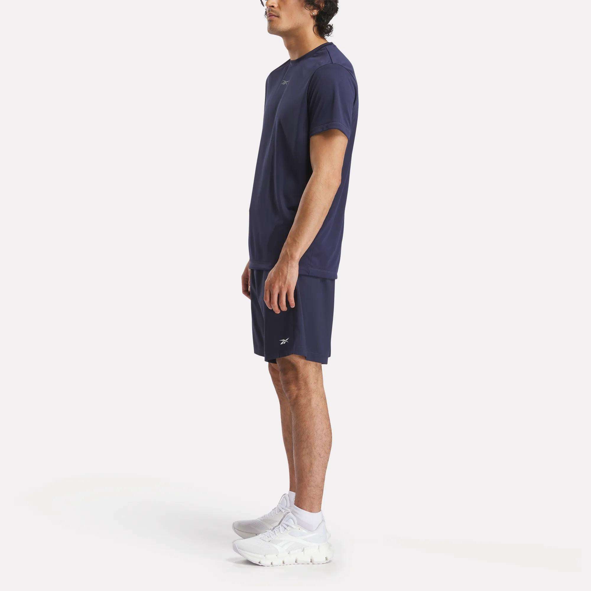 A man stands in profile wearing a navy blue athletic t-shirt, Reebok Men's Workout Ready Woven Shorts, and white sneakers against a plain white background.