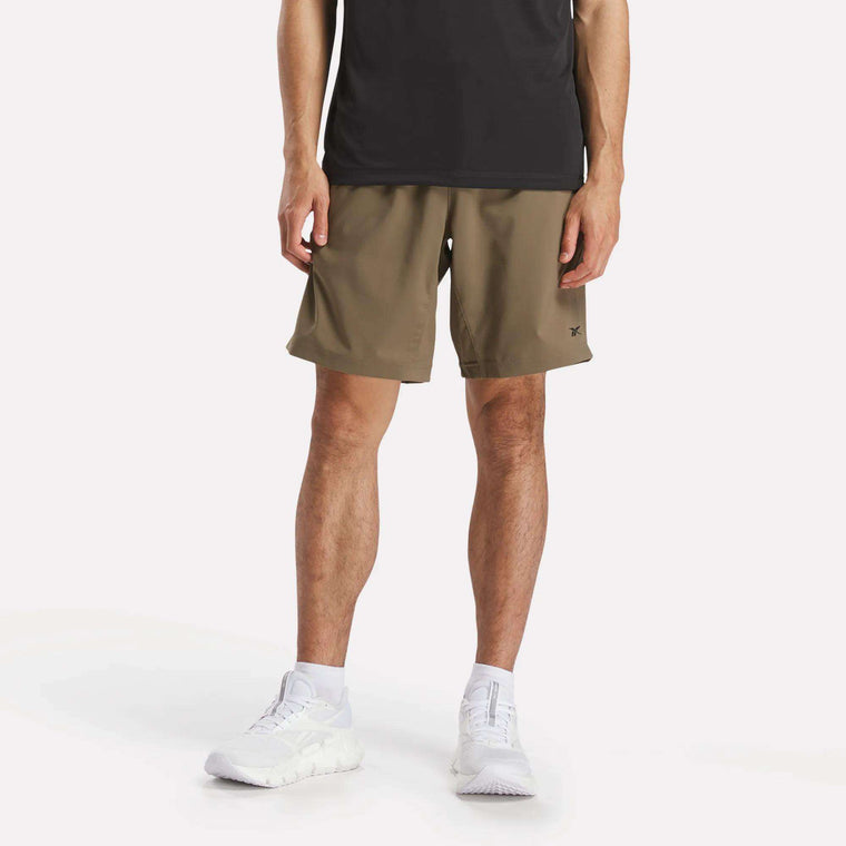 A person stands against a plain background, wearing a black t-shirt, Reebok Men's Workout Ready Woven Shorts in olive green, white socks, and white sneakers. The upper body is partially out of frame.