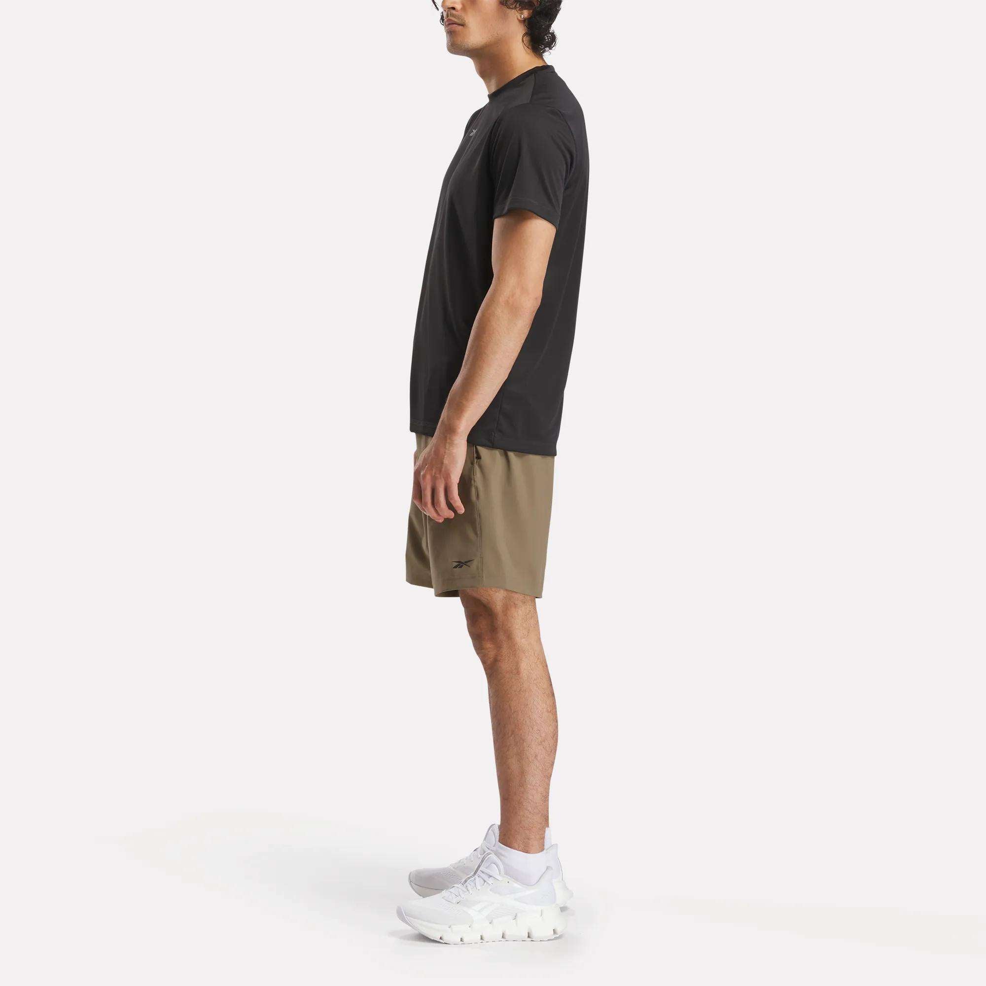 A man stands in profile against a plain background, wearing a black T-shirt, Reebok Men's Workout Ready Woven Shorts in khaki, and white athletic shoes.