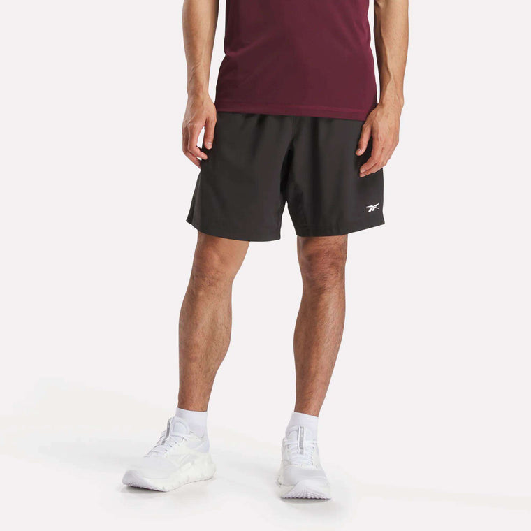 A person models the Reebok Men's Workout Ready Woven Shorts, shown from the waist down against a plain white background, paired with a burgundy T-shirt and white sneakers.