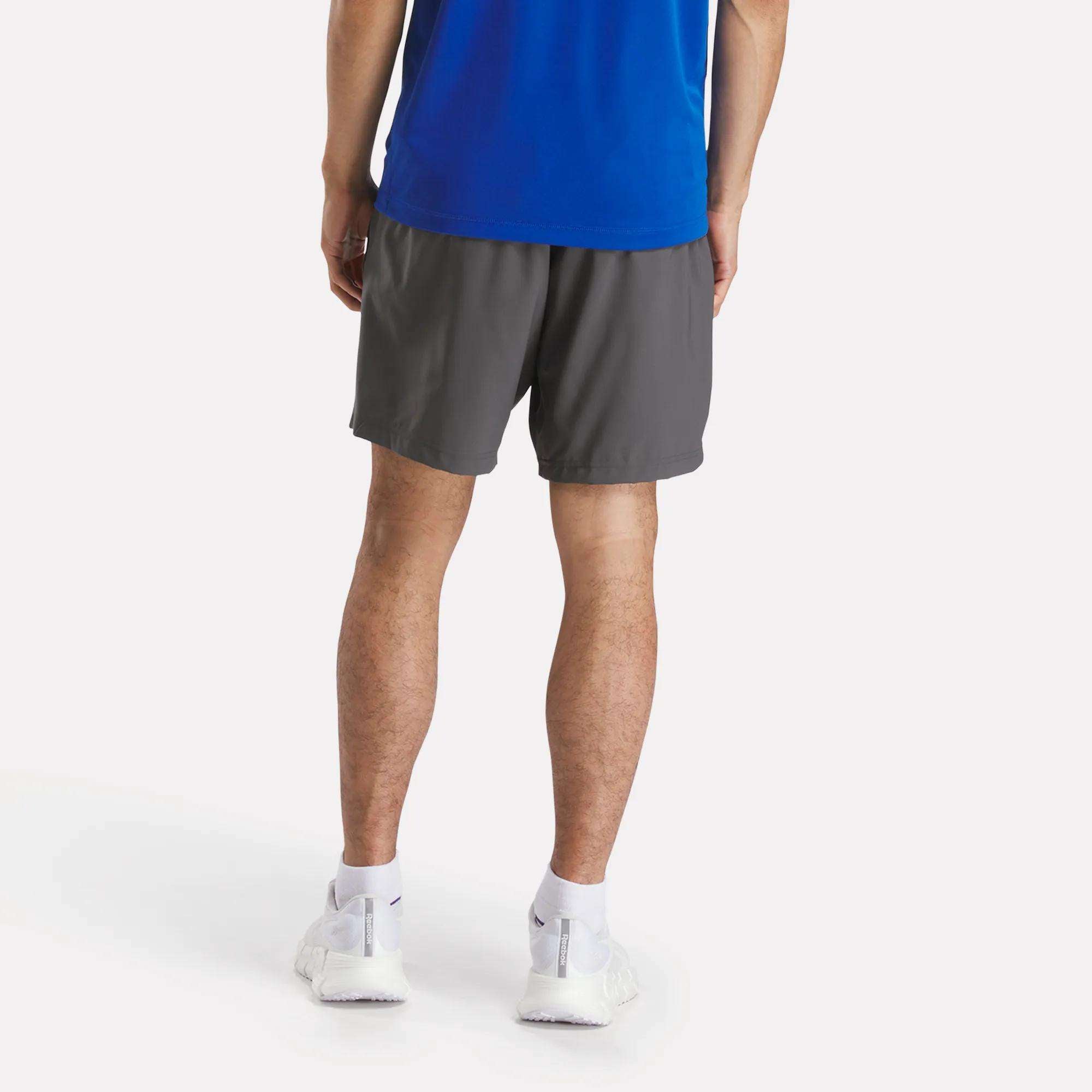 A person stands facing away, wearing a blue shirt, white socks, white sneakers, and gray Men's Workout Ready Woven Shorts by Reebok against a plain light background.