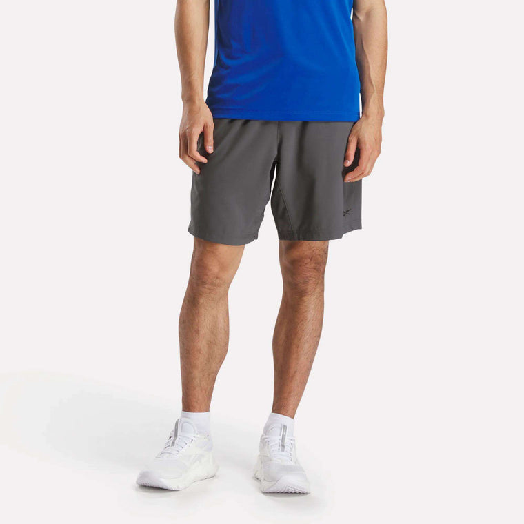 A person, visible from the waist down, stands against a white background with hands at their sides, wearing gray Men's Workout Ready Woven Shorts by Reebok, a blue shirt, and white sneakers.