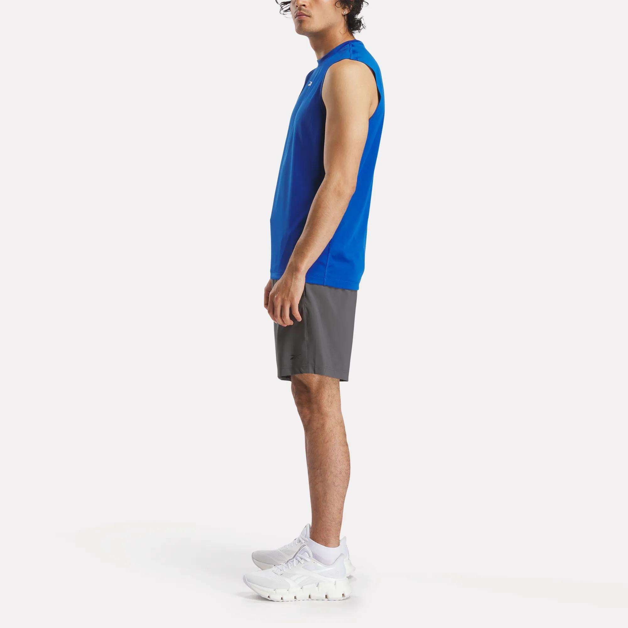 A man stands in profile wearing a sleeveless blue athletic shirt, Reebok Men's Workout Ready Woven Shorts, and white sneakers against a plain white background.