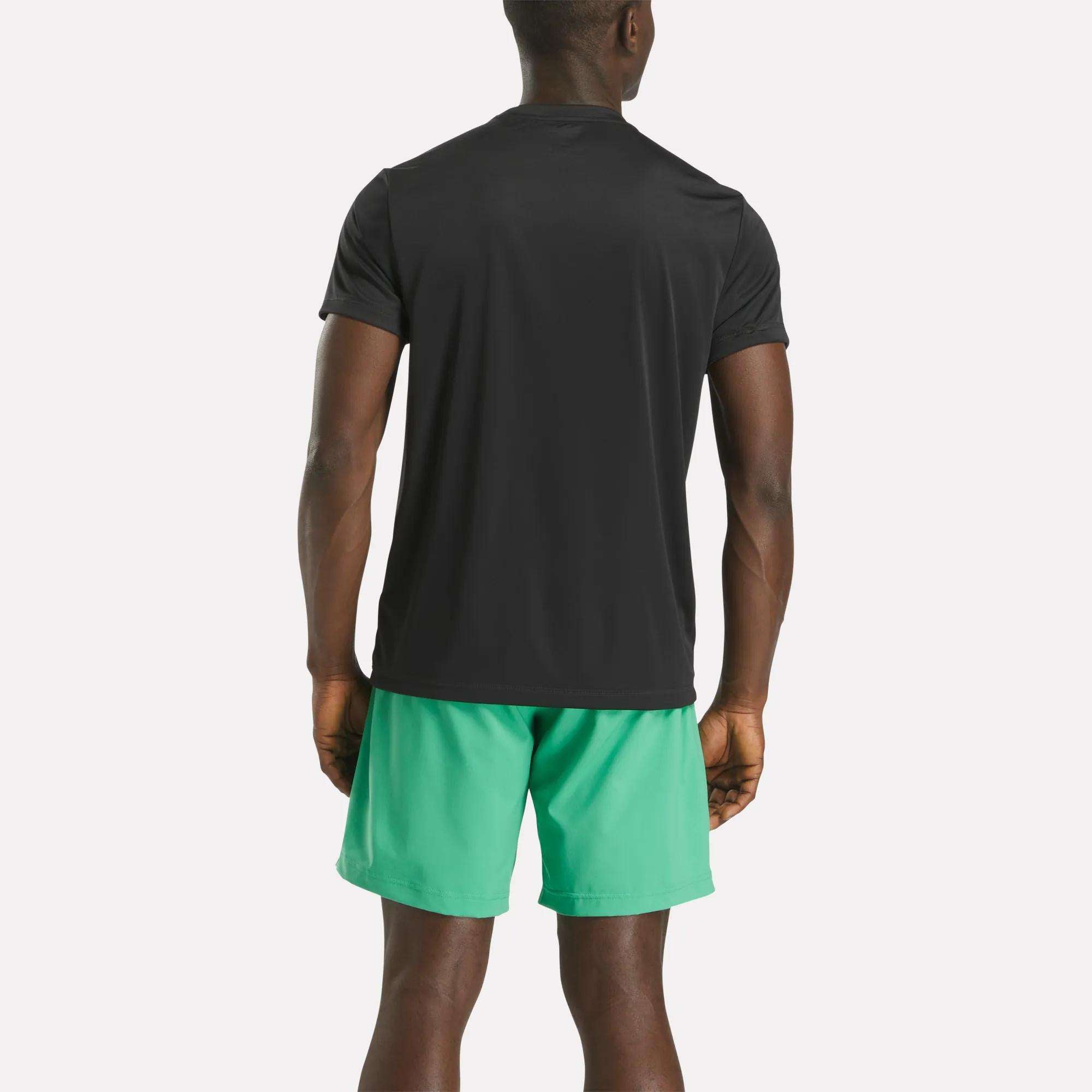 A person stands facing away from the camera, wearing a black Reebok Men's ID Train Short Sleeve Tech Tee and green athletic shorts against a plain white background.