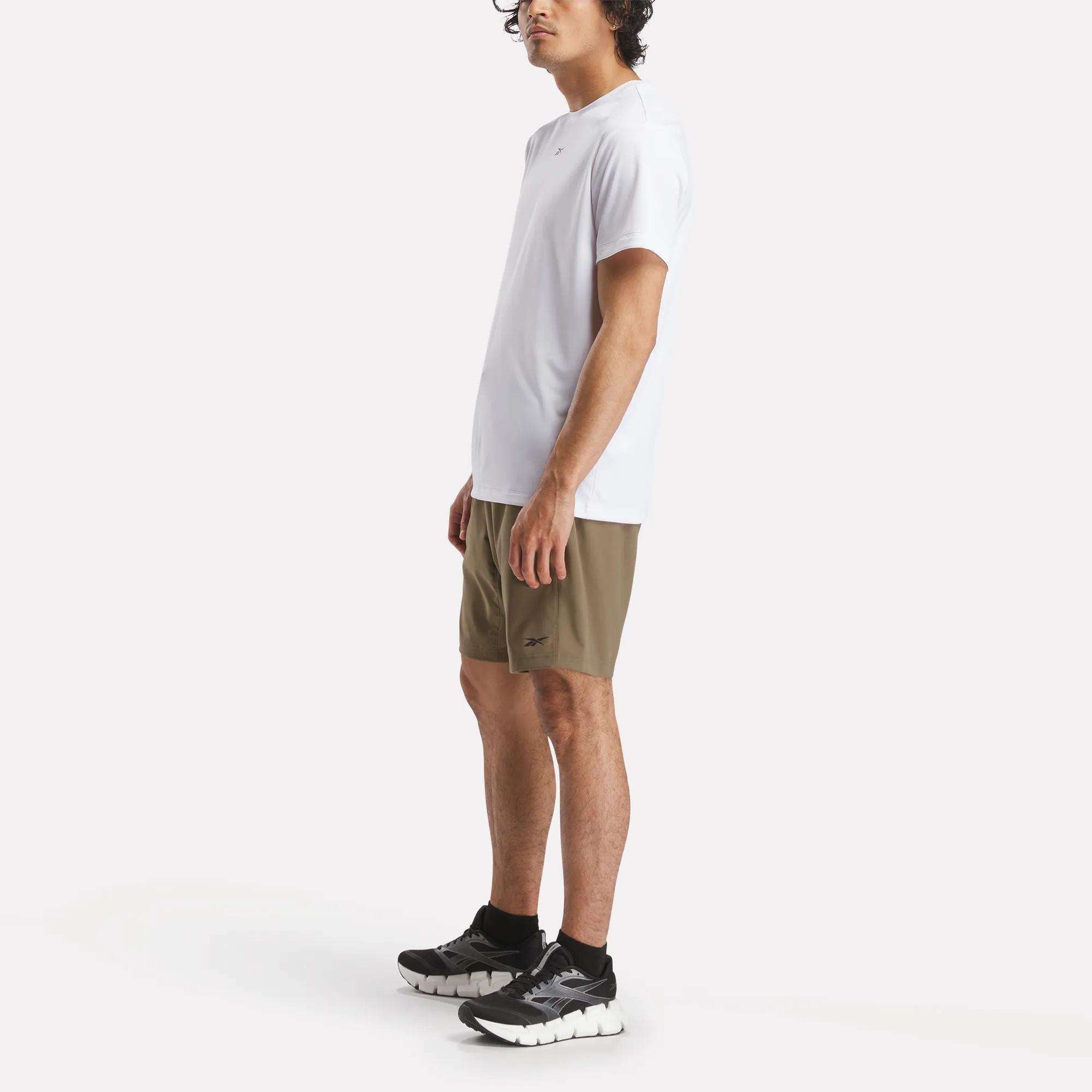 A man stands against a white background wearing the Reebok Men's ID Train Short Sleeve Tech Tee, olive green shorts, black socks, and black running shoes. He faces slightly sideways with his arms by his sides.