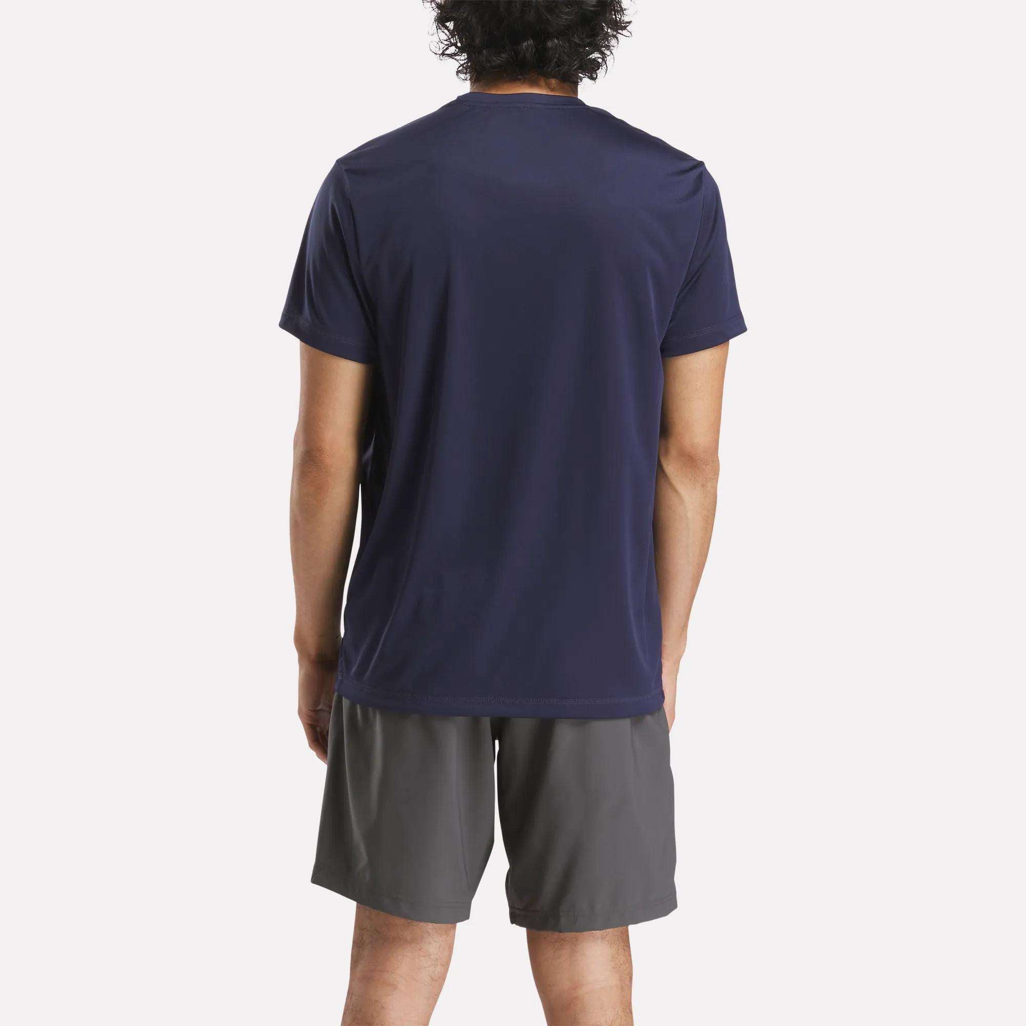 A person with short curly hair, seen from behind, wears a dark navy Reebok Men's ID Train Short Sleeve Tech Tee and gray athletic shorts against a plain white background.
