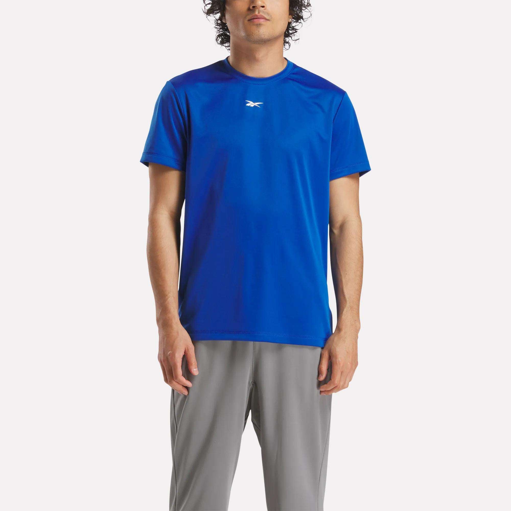 A man wears the Reebok Men's ID Train Short Sleeve Tech Tee in bright blue, featuring a small white chest logo and moisture-wicking RBK-DRY fabric, paired with light gray pants against a plain light backdrop.