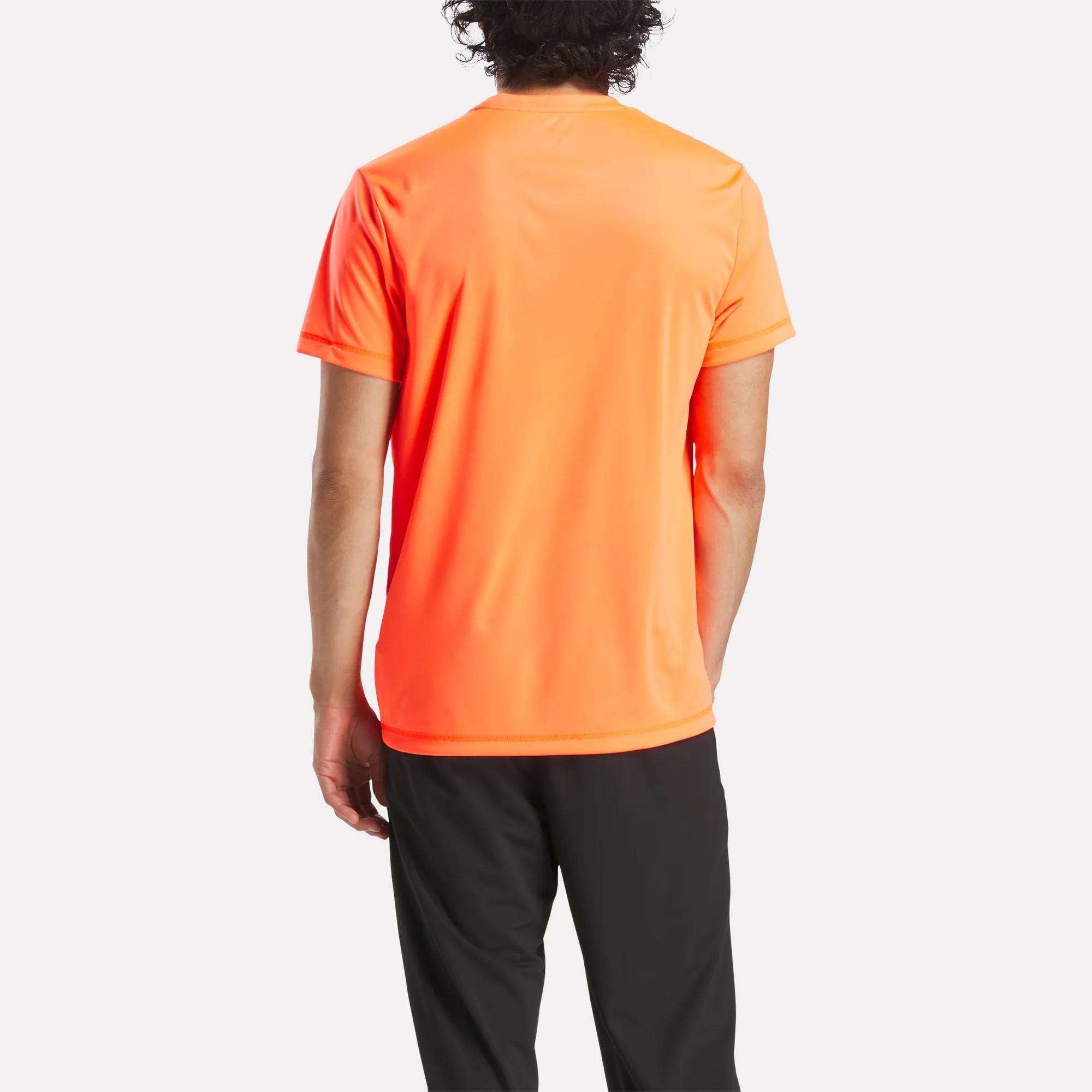 A person with curly hair stands with their back to the camera, wearing a bright orange Reebok Men's ID Train Short Sleeve Tech Tee and black pants against a plain white background.