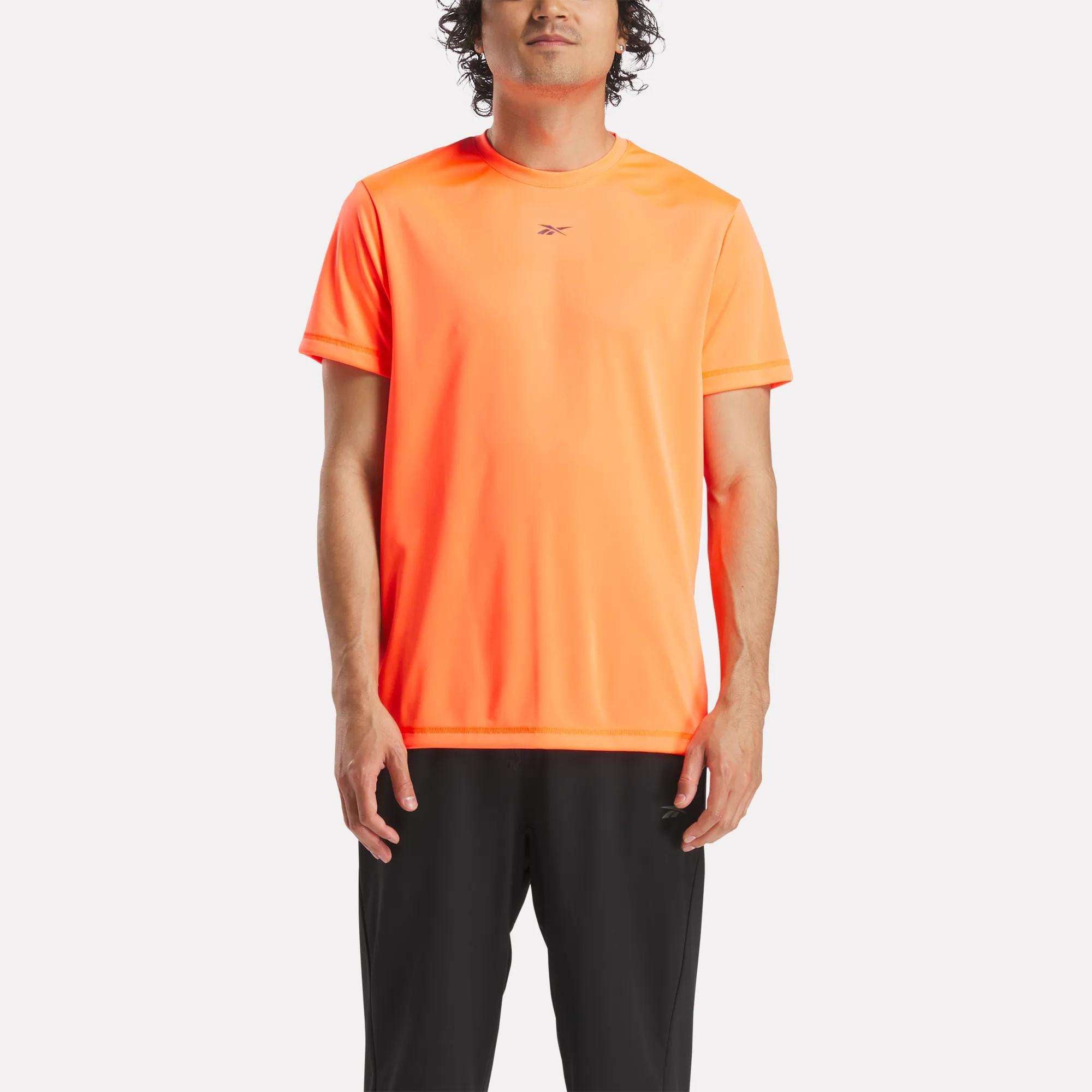 A man wears a bright orange Reebok Men's ID Train Short Sleeve Tech Tee and black pants, standing facing forward against a plain white background.