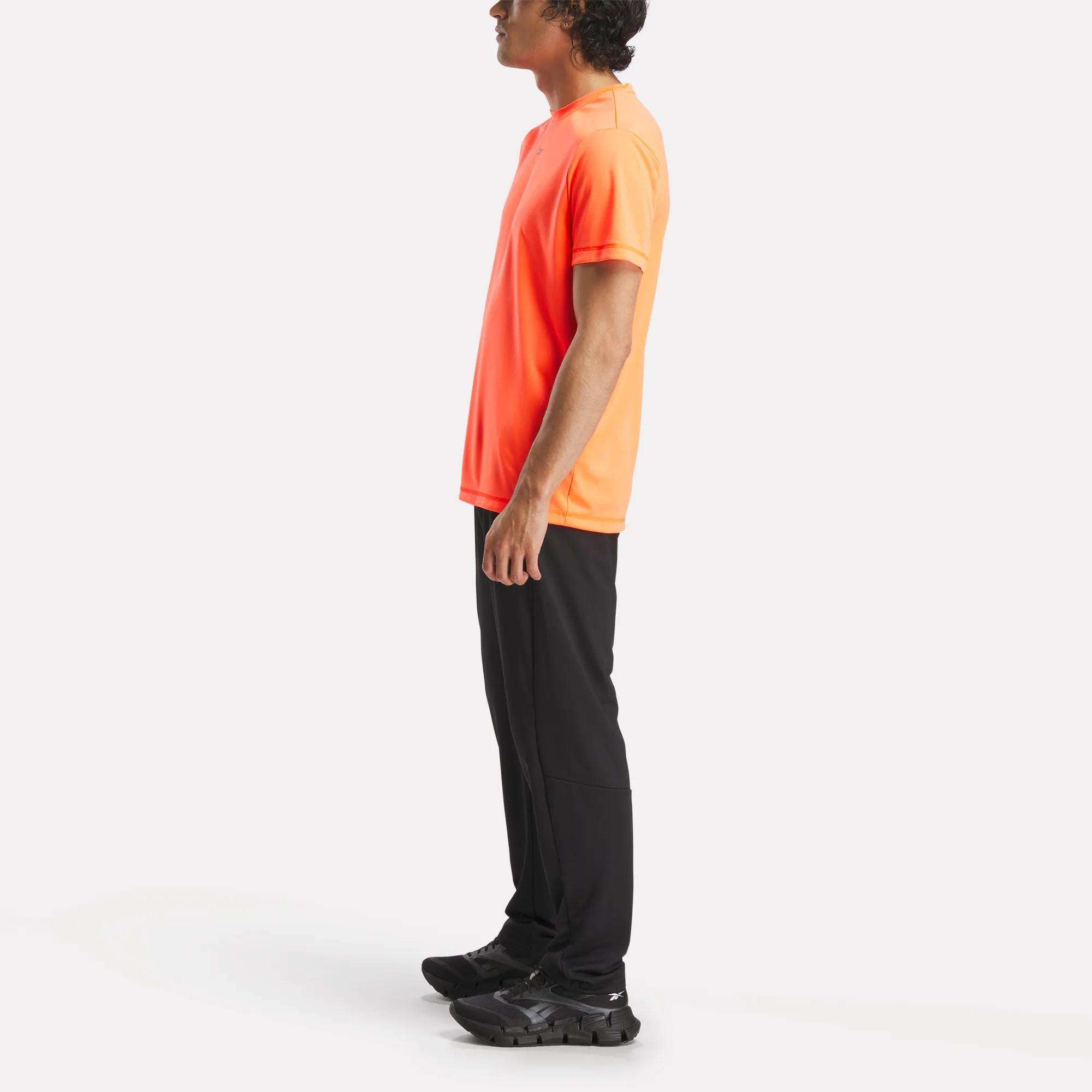 A man in profile wears a Reebok Men's ID Train Short Sleeve Tech Tee in orange, paired with black pants and sneakers, set against a plain white background.