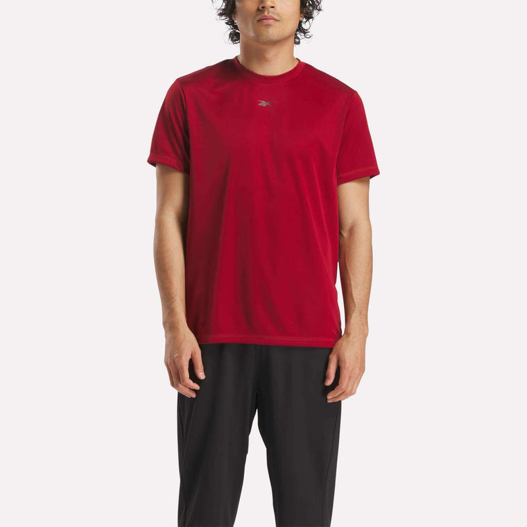 A man stands facing forward against a plain white background wearing a Reebok Men's ID Train Short Sleeve Tech Tee in red and black pants.