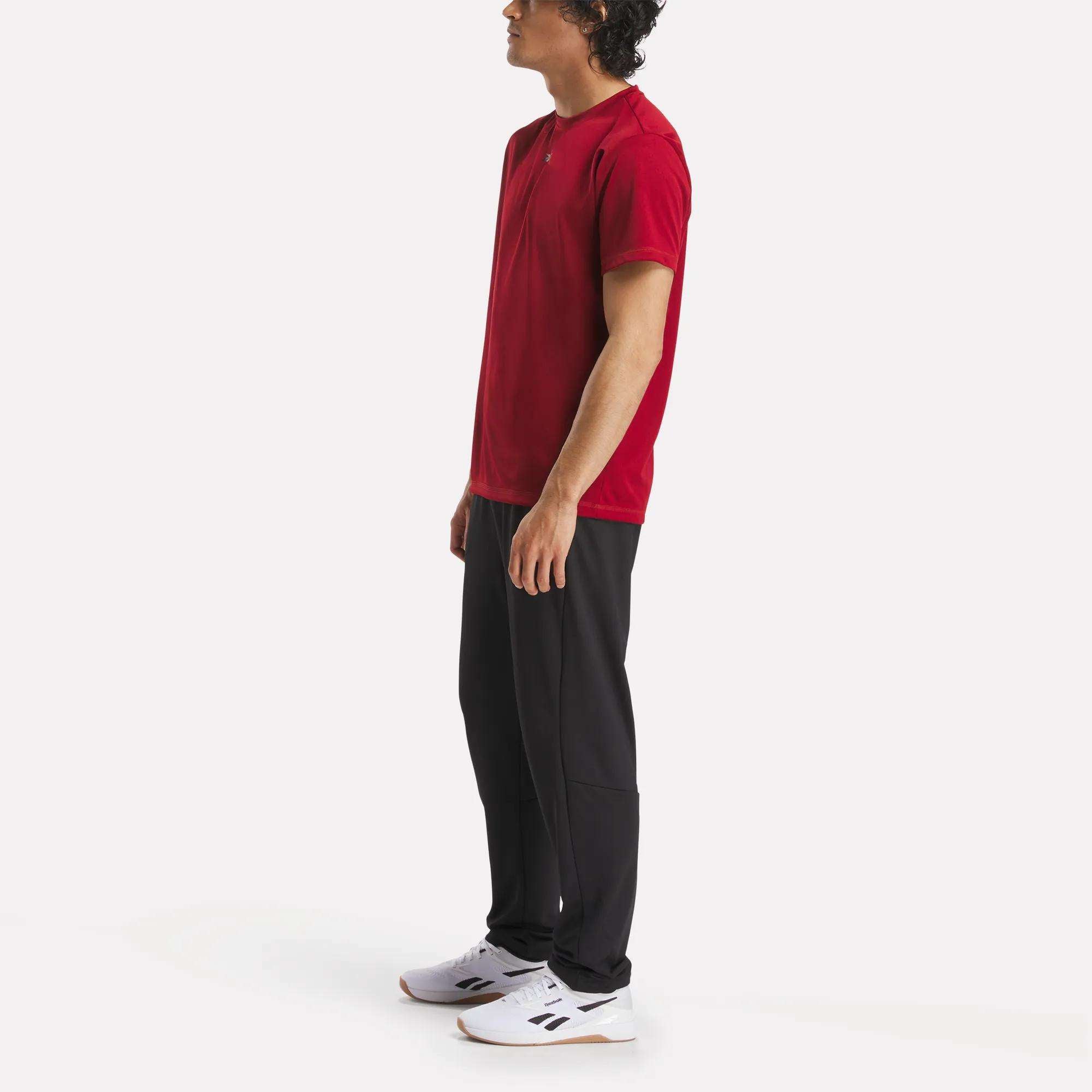 A person stands sideways wearing a red Reebok Men's ID Train Short Sleeve Tech Tee, black pants, and white sneakers against a plain white background.