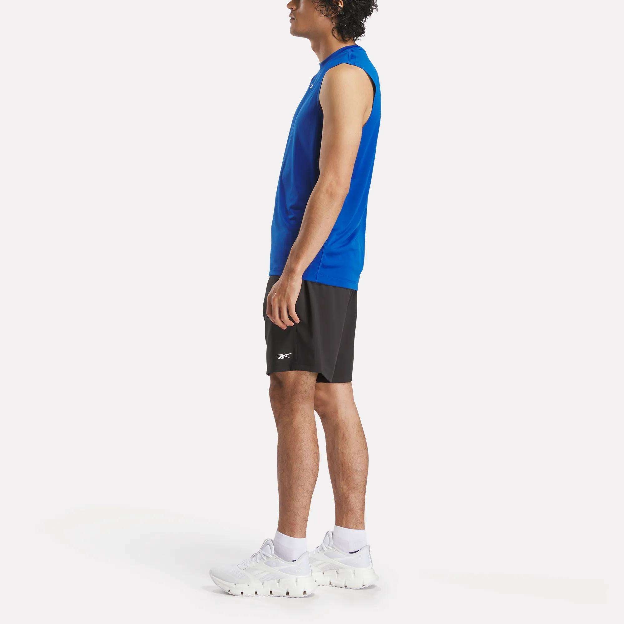 A man stands in profile wearing the Reebok Men's Train Sleeveless Tech Tee, black athletic shorts, and white sneakers against a plain white background.