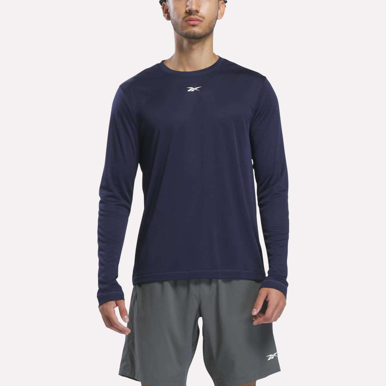 A person wearing a Reebok Men’s Long Sleeve Tech T-Shirt in Vector Navy and gray Reebok shorts stands facing forward against a plain white background.