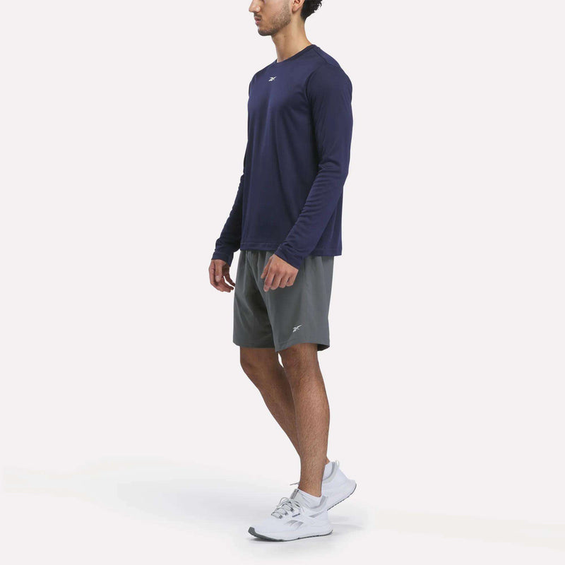 A man stands in profile on a plain white background wearing a navy Reebok Men’s Long Sleeve Tech T-Shirt, gray athletic shorts, and white sneakers.
