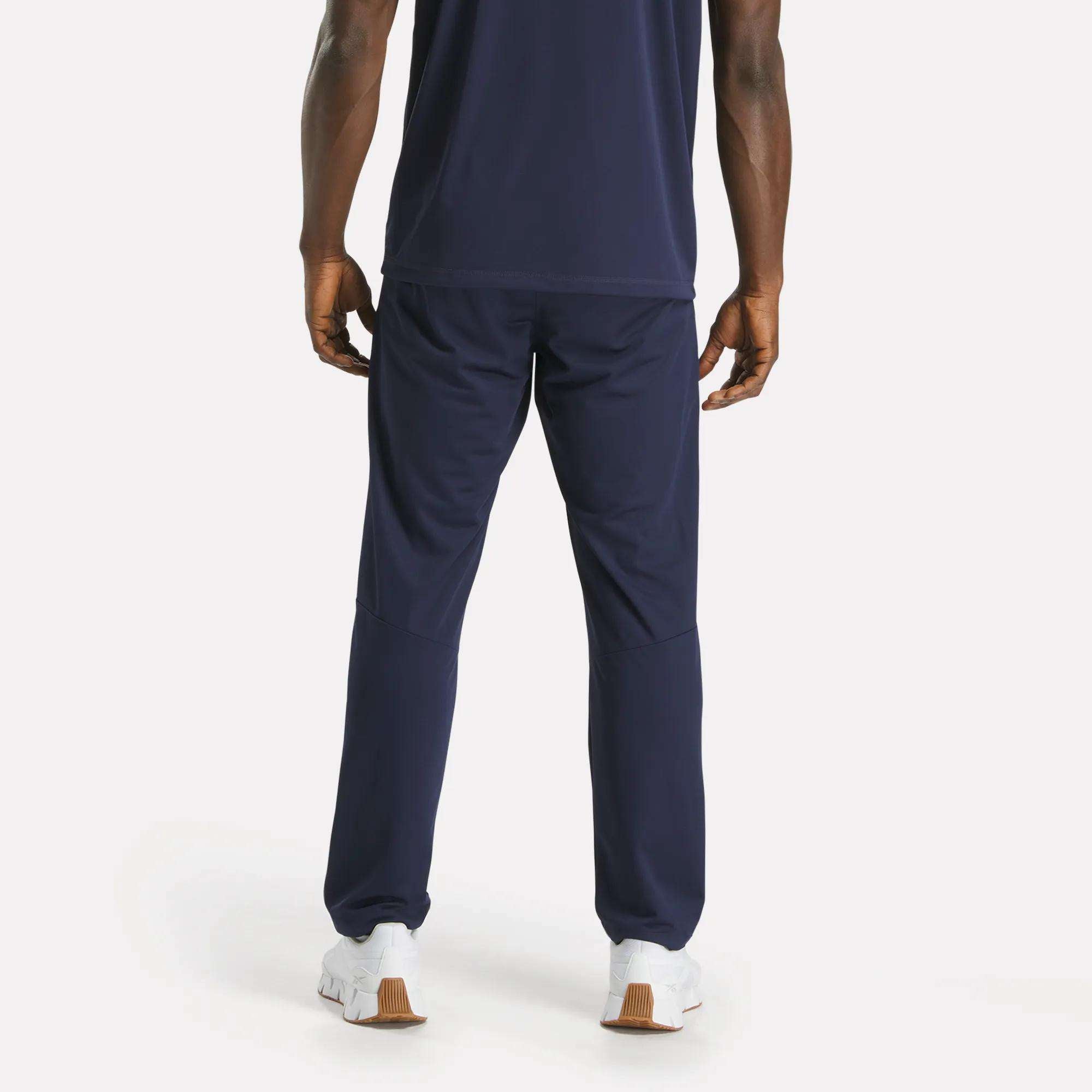 A man faces away, wearing a navy blue T-shirt and Reebok Men's Train Knit Pants with white sneakers on a plain light background.