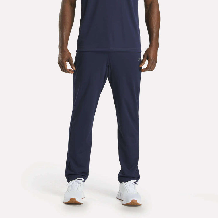 A person in navy blue Reebok Men's Train Knit Pants stands against a plain white background, paired with a matching T-shirt and white sneakers. The image is cropped just below the shoulders.