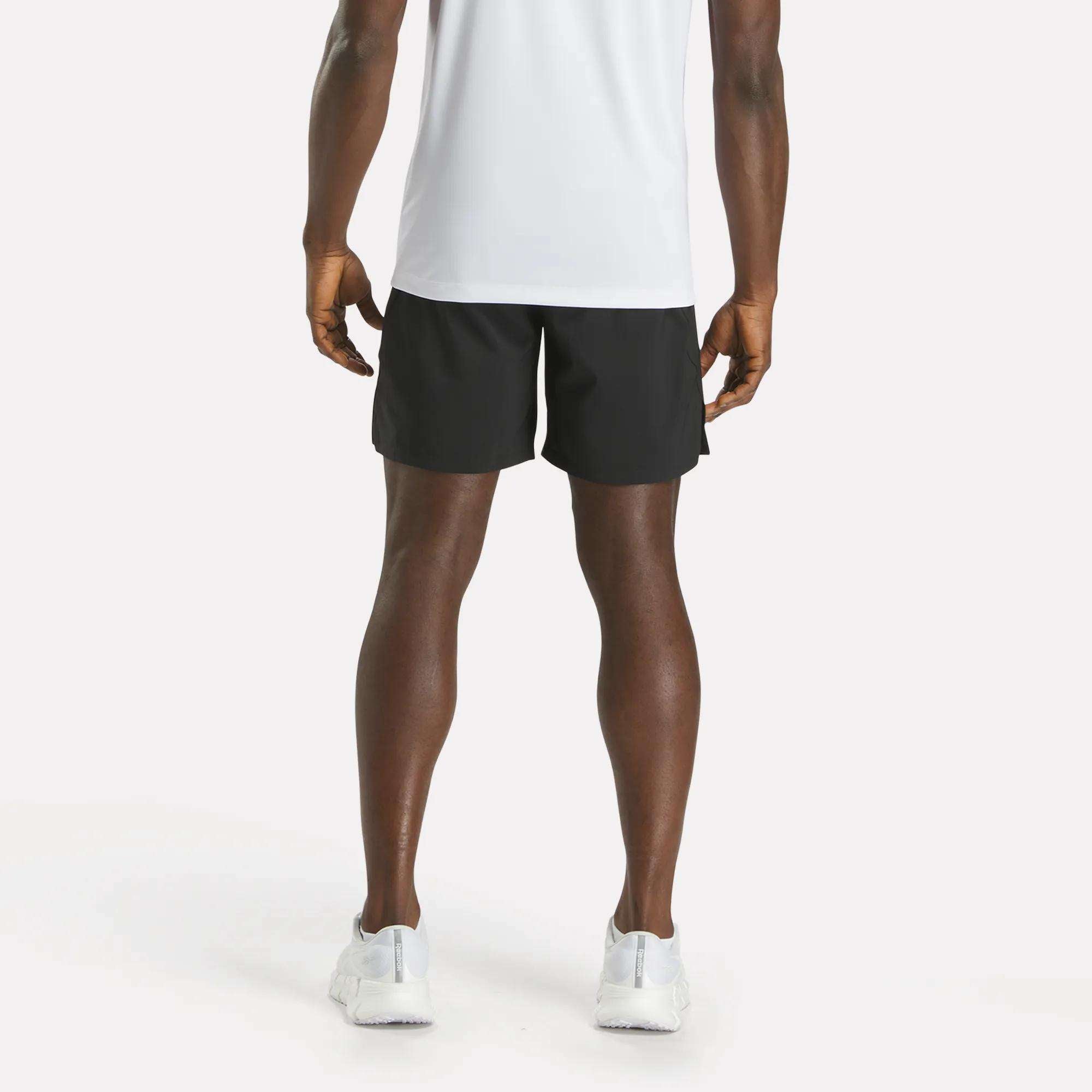 A person stands facing away from the camera, wearing a white T-shirt, Reebok Men's Athlete Speed Shorts in black, and white sneakers against a plain light background.