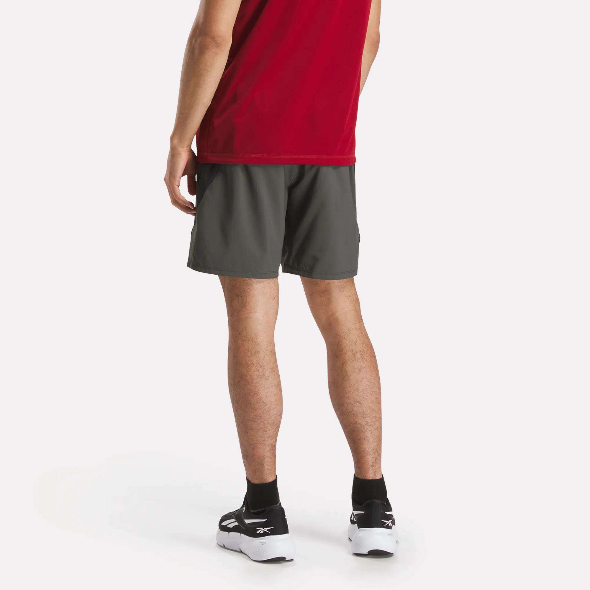 A person stands facing away, wearing a red shirt, Reebok Men's Athlete Speed Shorts in dark gray with RBK-DRY technology, and black and white sneakers against a plain white background.