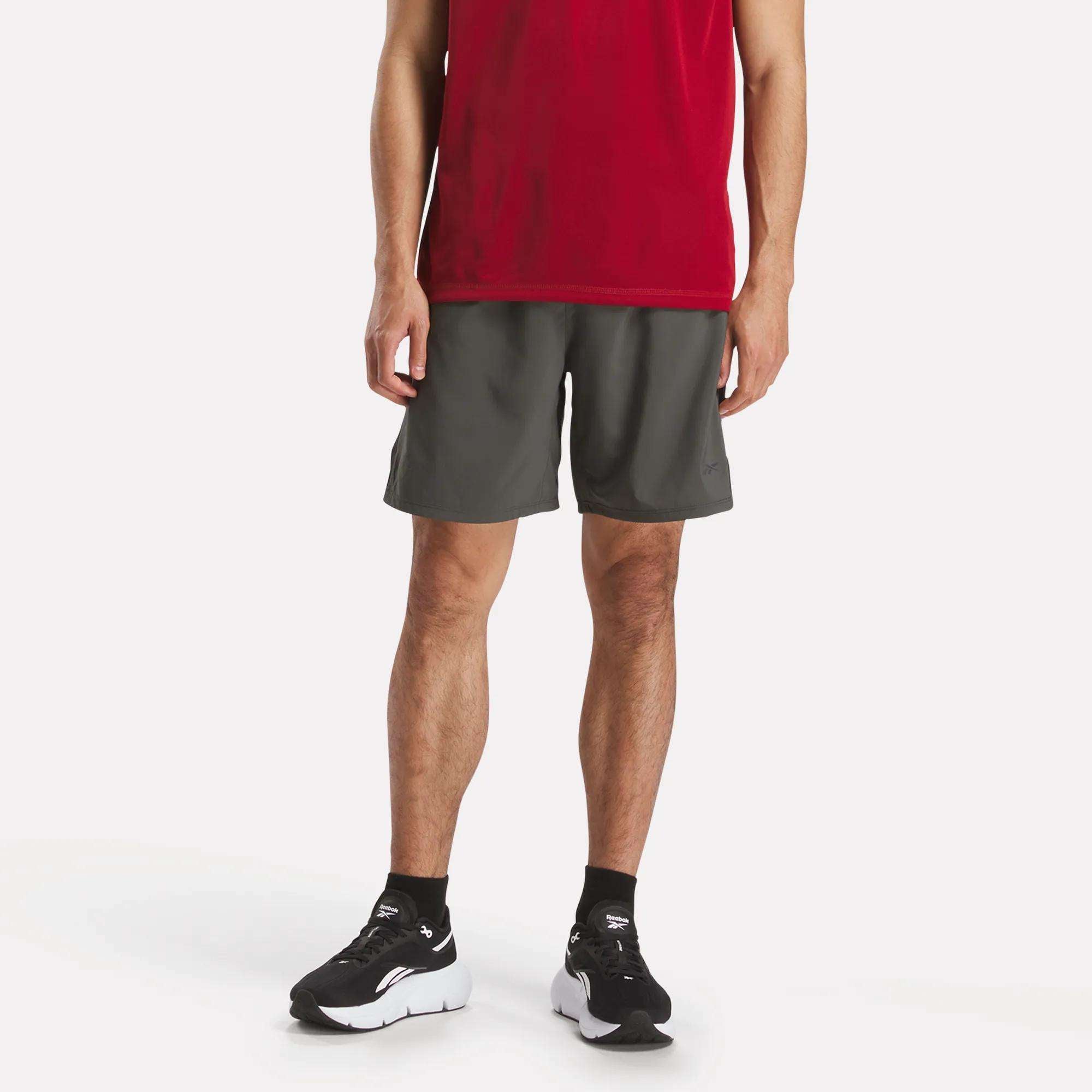 A person in a red shirt, Reebok Men’s Athlete Speed Shorts in dark gray, black socks, and black and white sneakers stands against a plain white background. The face is not visible.