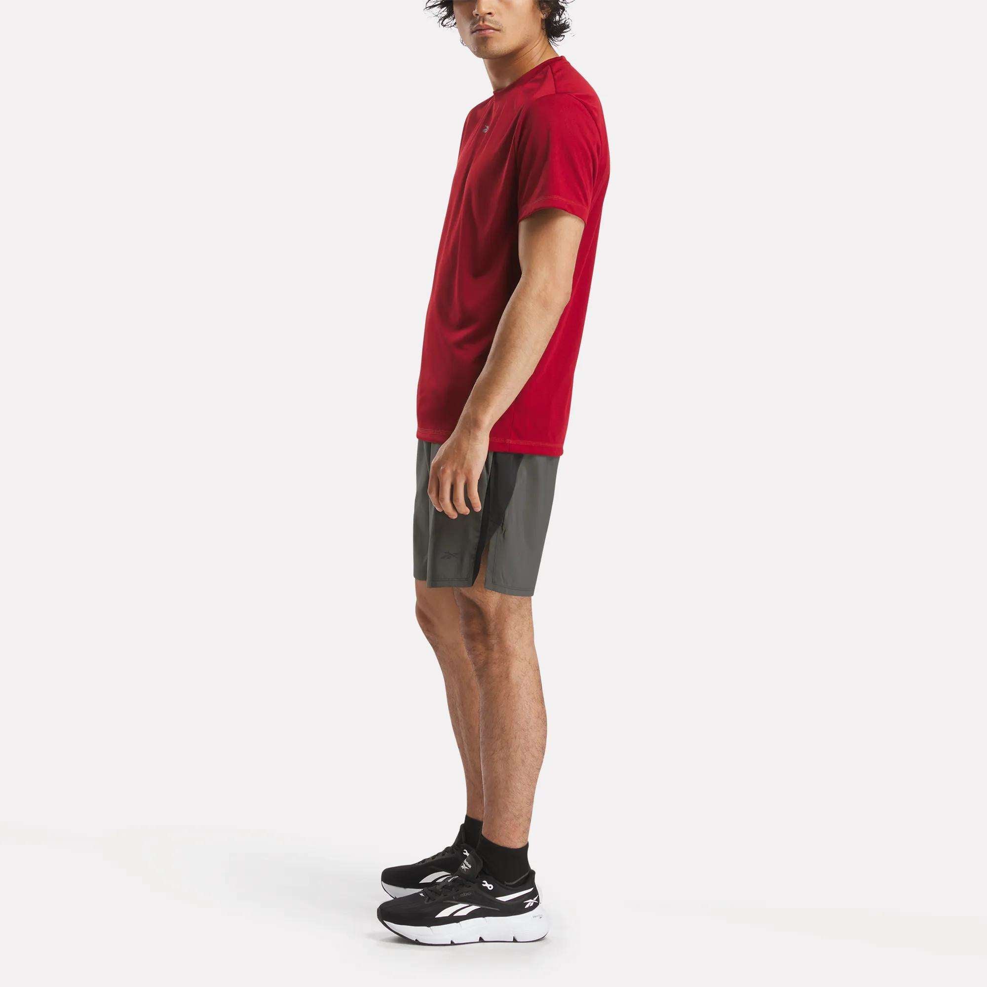 A person in a red T-shirt, black socks, black athletic shoes, and dark gray Reebok Men's Athlete Speed Shorts with RBK-DRY technology stands facing left against a plain white background.