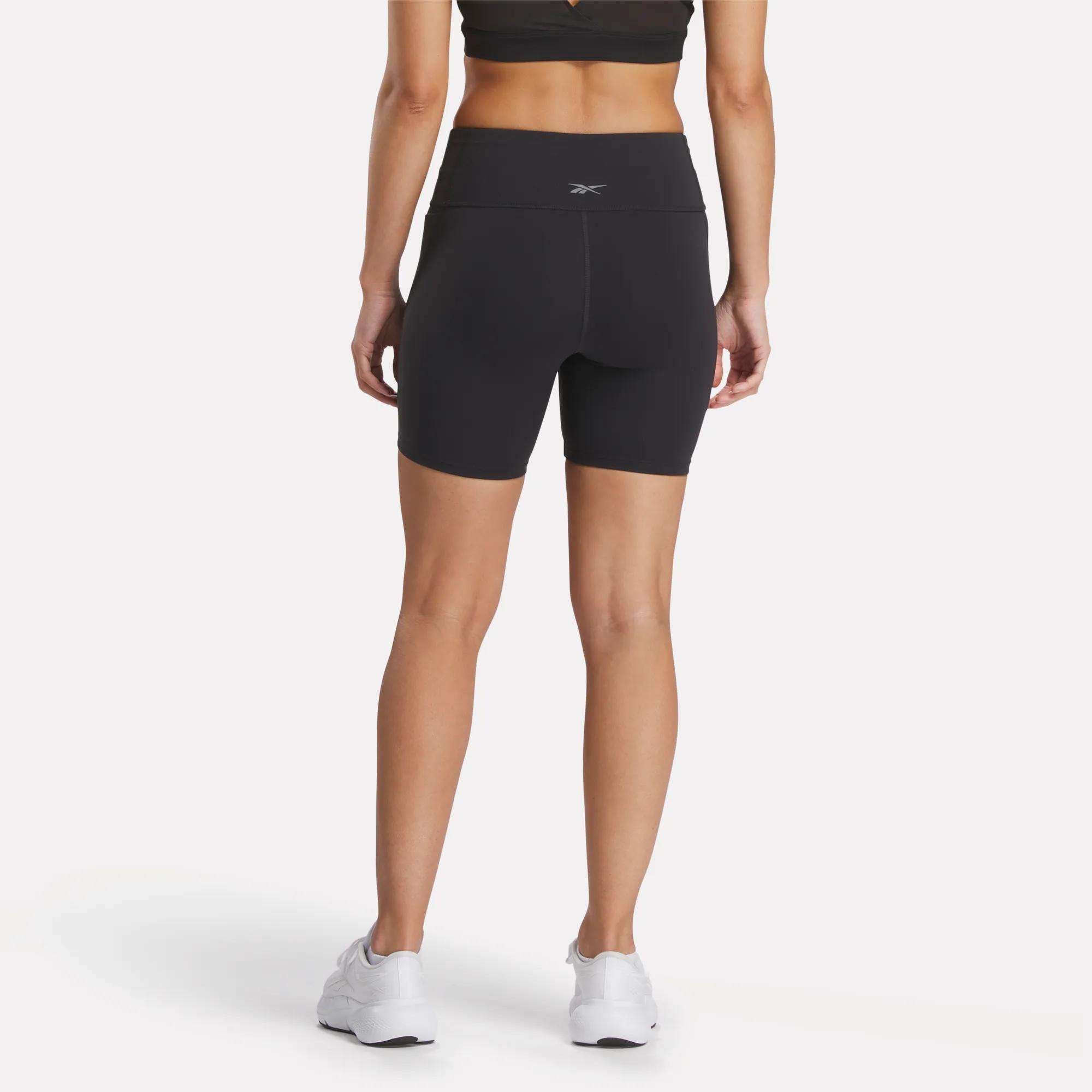 A person wearing Reebok Women's Lux High-Rise Bike Shorts in black, a matching black sports bra, and white sneakers stands facing away against a plain white background.