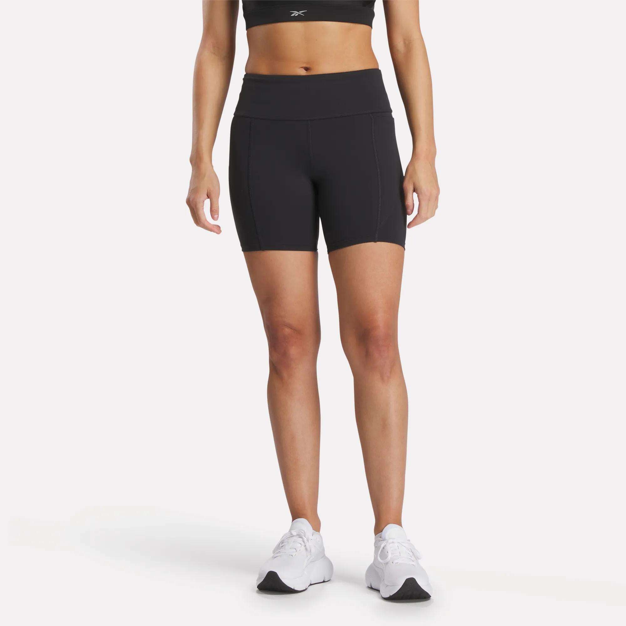 A woman, shown from the shoulders down, wears a black RBK-DRY sports bra, Reebok Women's Lux High-Rise Bike Shorts, and white sneakers against a plain white background.