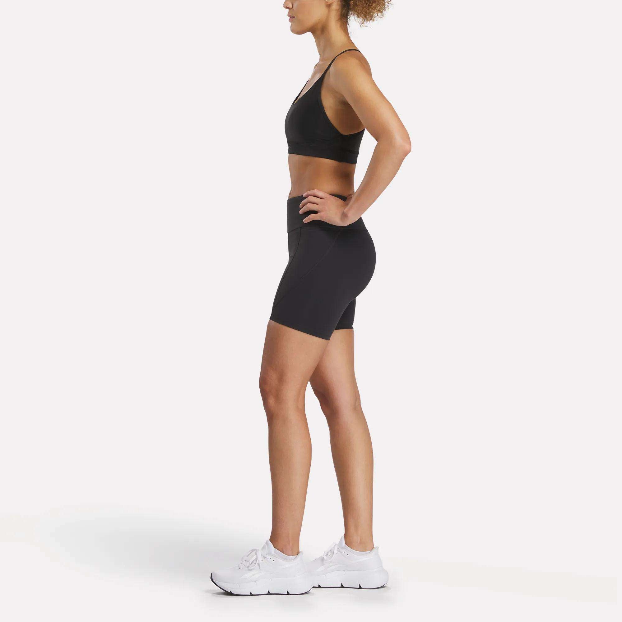 A woman in profile poses against a white background, wearing a black sports bra, Reebok Women's Lux High-Rise Bike Shorts featuring RBK-DRY technology, and white sneakers. Her arms are bent with her hands on her hips.