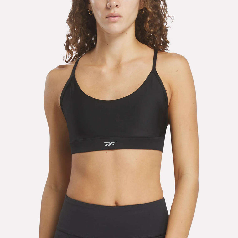 A woman with curly hair wears the Reebok Women's Lux Strappy Sports Bra, featuring thin straps and a small logo, against a plain light background.