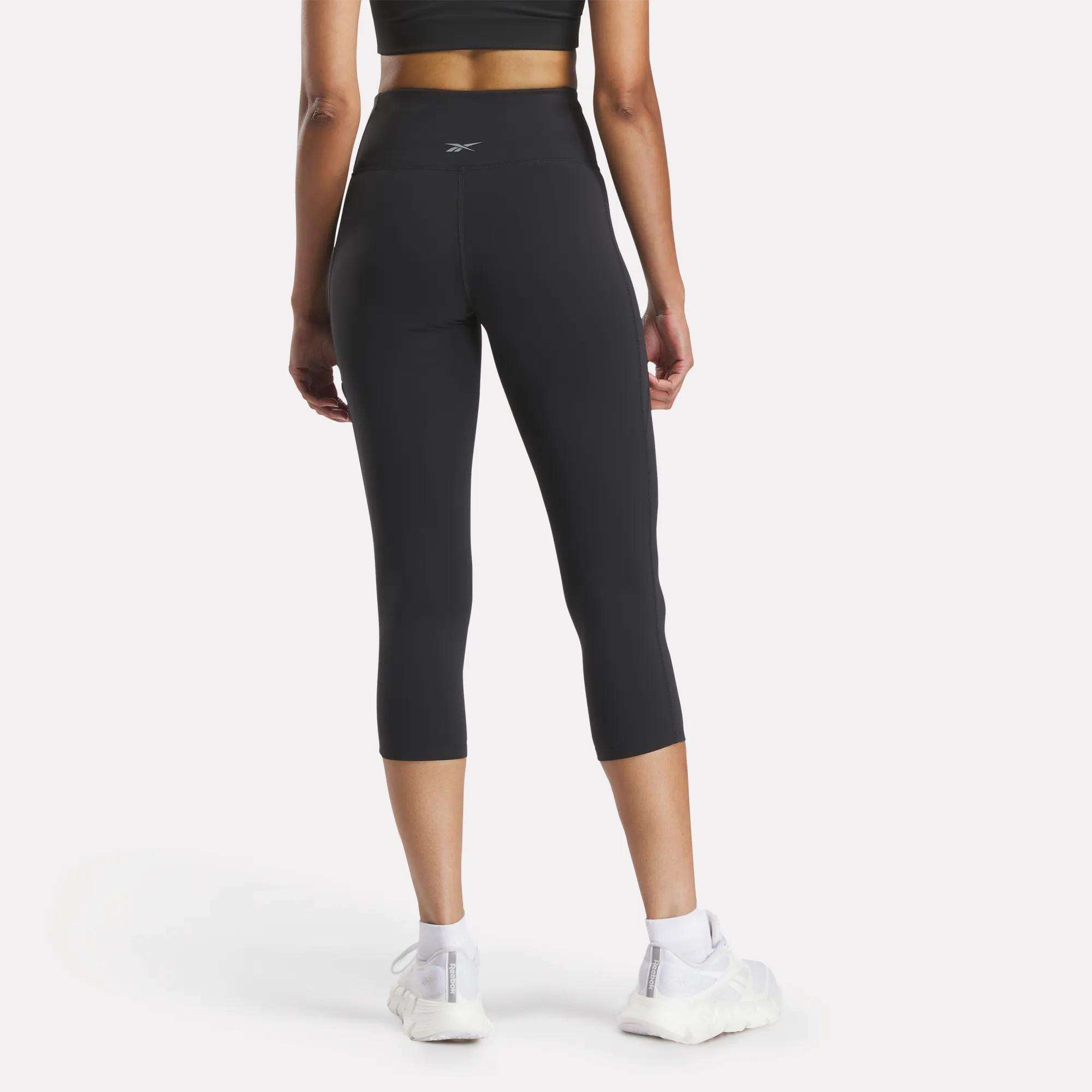 A woman stands facing away, wearing a black sports bra and Reebok Women's Lux 3/4 High-Rise Tights with a high-rise waistband, paired with white sneakers against a plain white background.