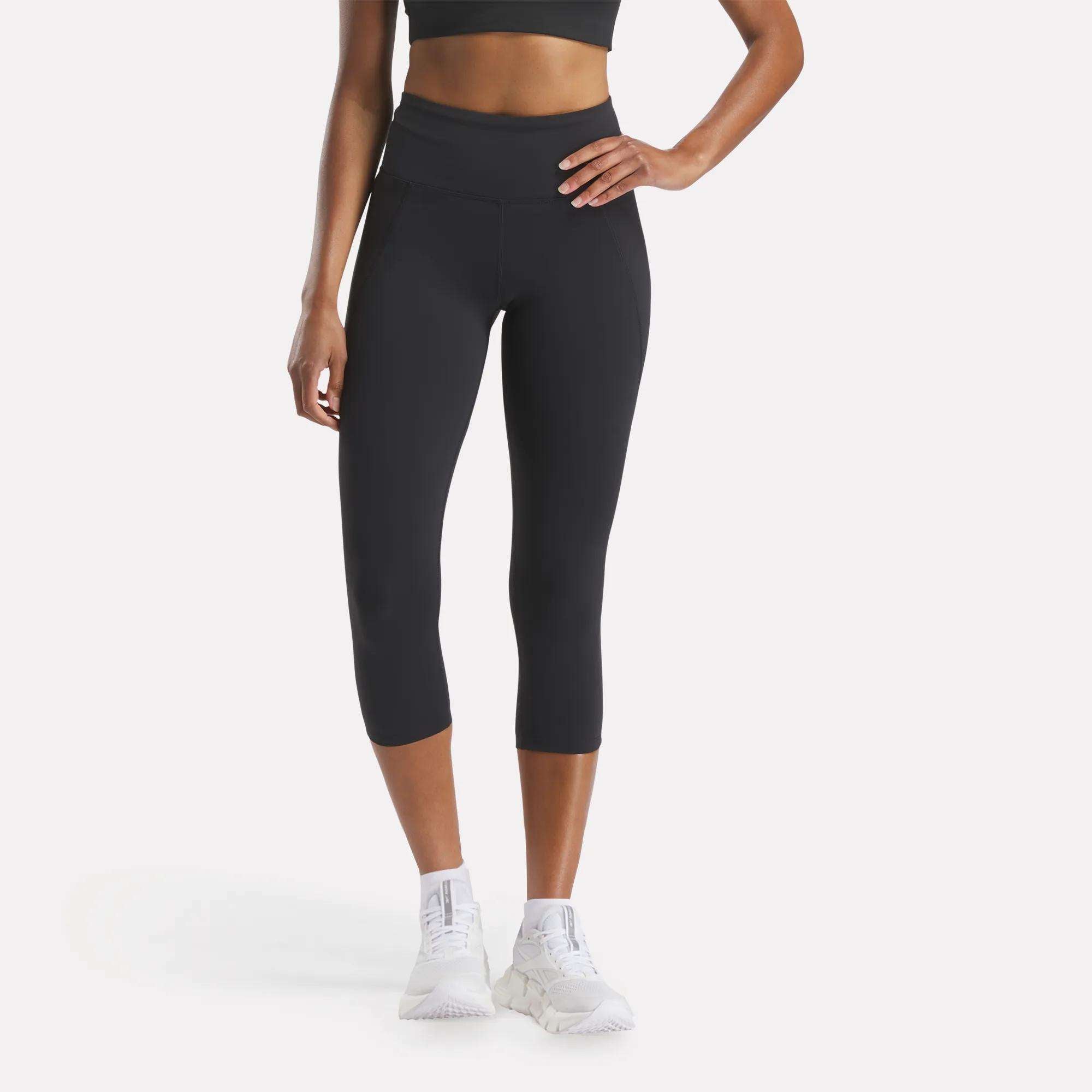 A woman in a black sports bra, Reebok Women's Lux 3/4 High-Rise Tights, and white sneakers stands with one hand on her hip against a plain white background.