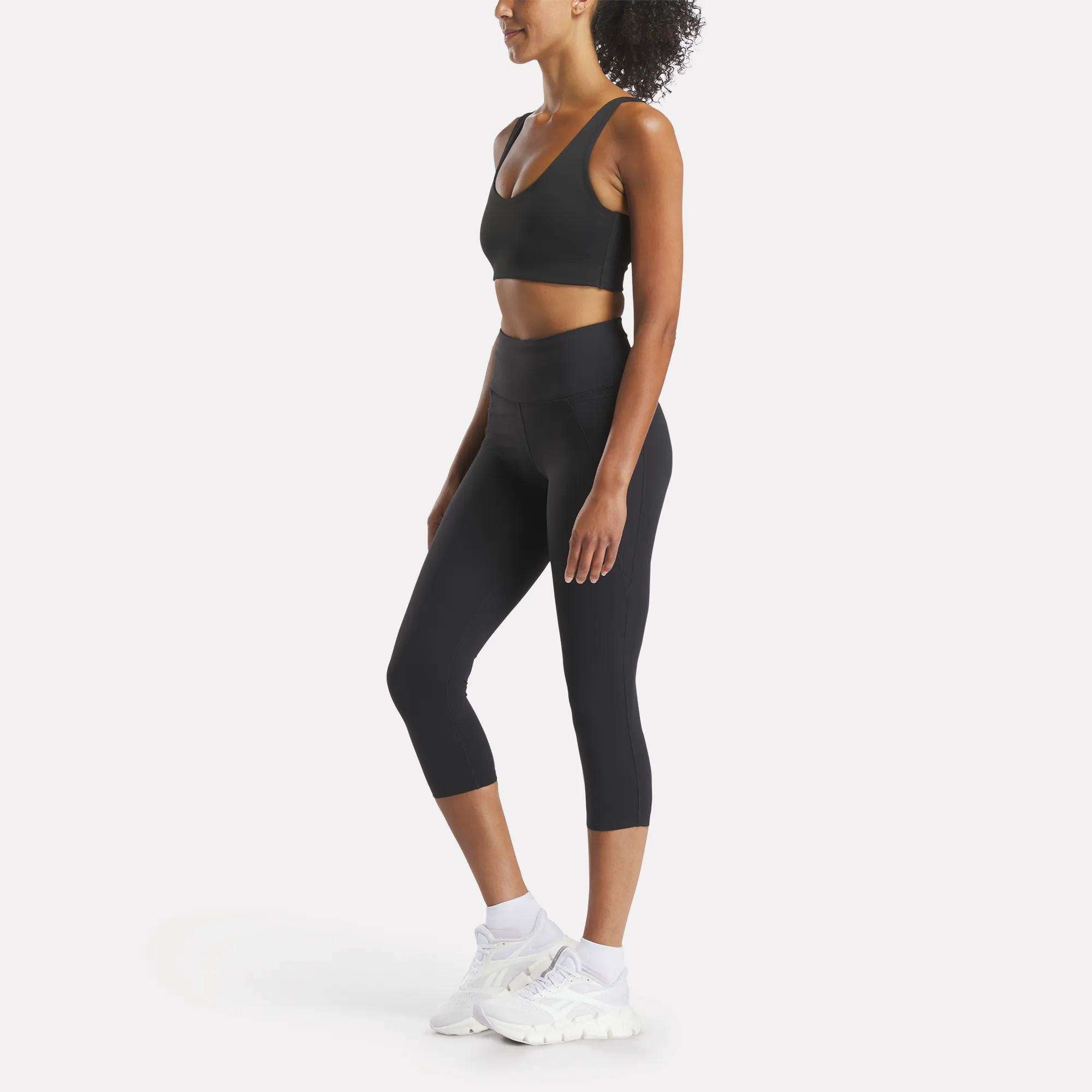 A woman stands in profile against a white background, wearing a black sports bra, Reebok Women's Lux 3/4 High-Rise Tights, white socks, and white sneakers.