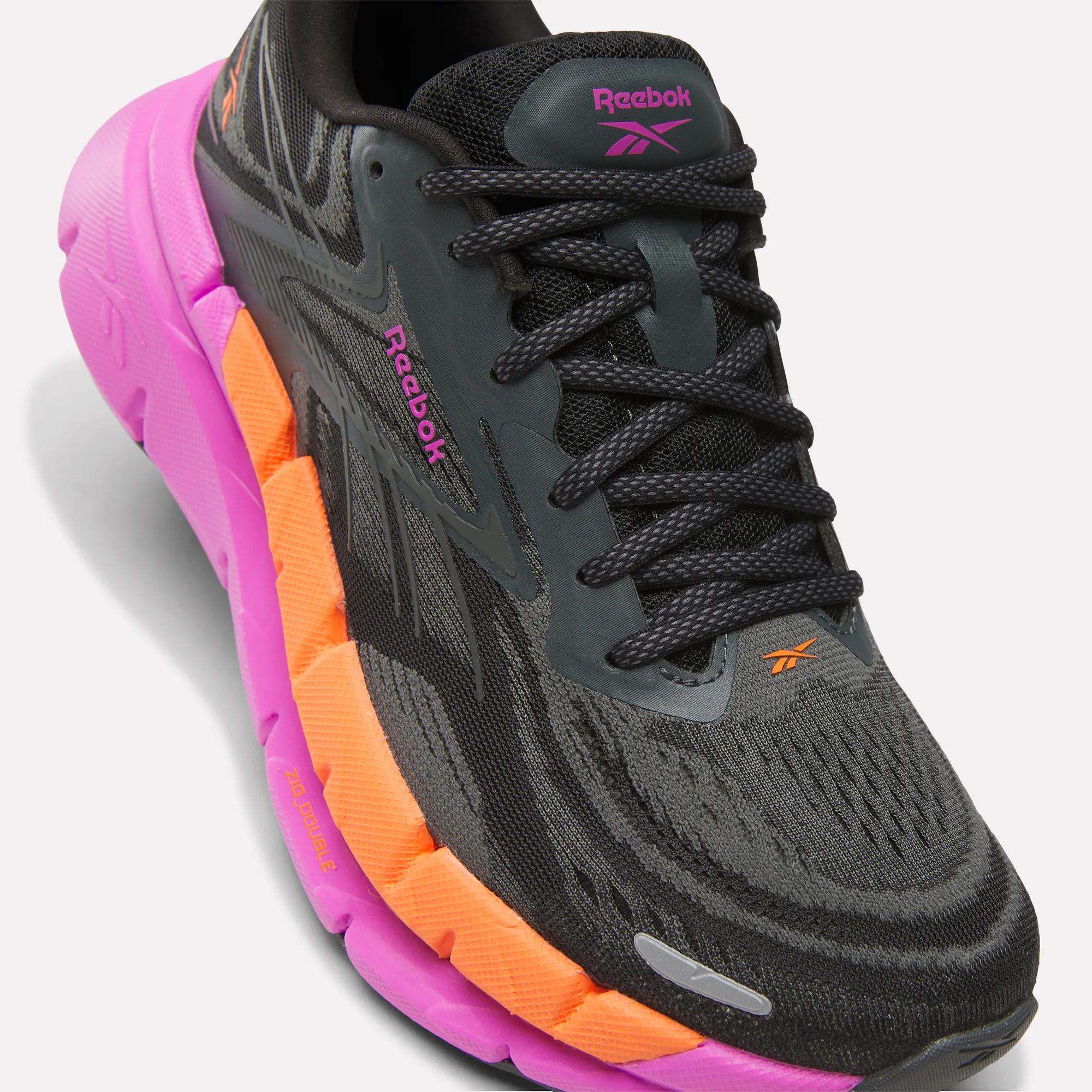 A close-up of the Reebok Women's FloatZig Double Running Shoe in black and gray with pink and orange accents, black laces, and visible Reebok logos, designed for max cushioning, set against a white background.