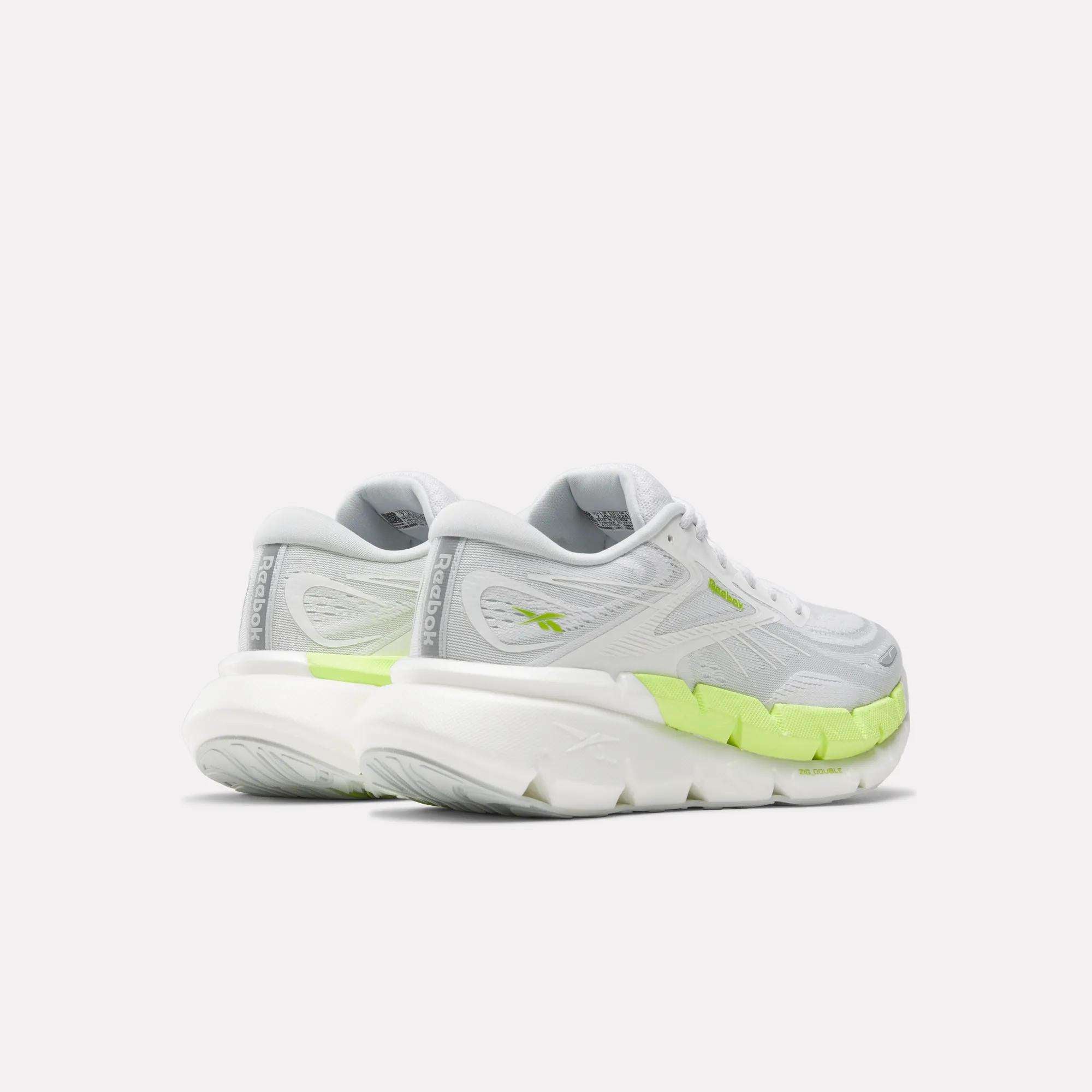 The Women's FloatZig Double Running Shoes by Reebok feature chunky soles and neon green accents on the sides and heel, offering maximum cushioning for running. Shown in white on a plain background.