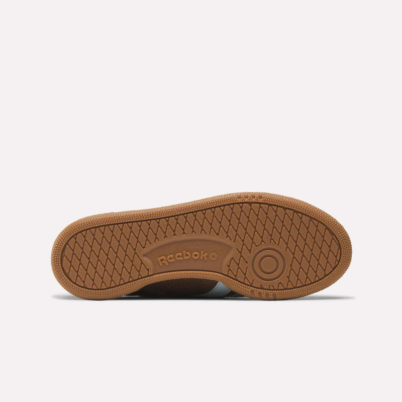 The image shows the gum rubber sole of the Reebok Women's Club C Grounds UK Shoes, featuring a herringbone tread pattern, a circular design near the toe area, and "Reebok" embossed in the center.