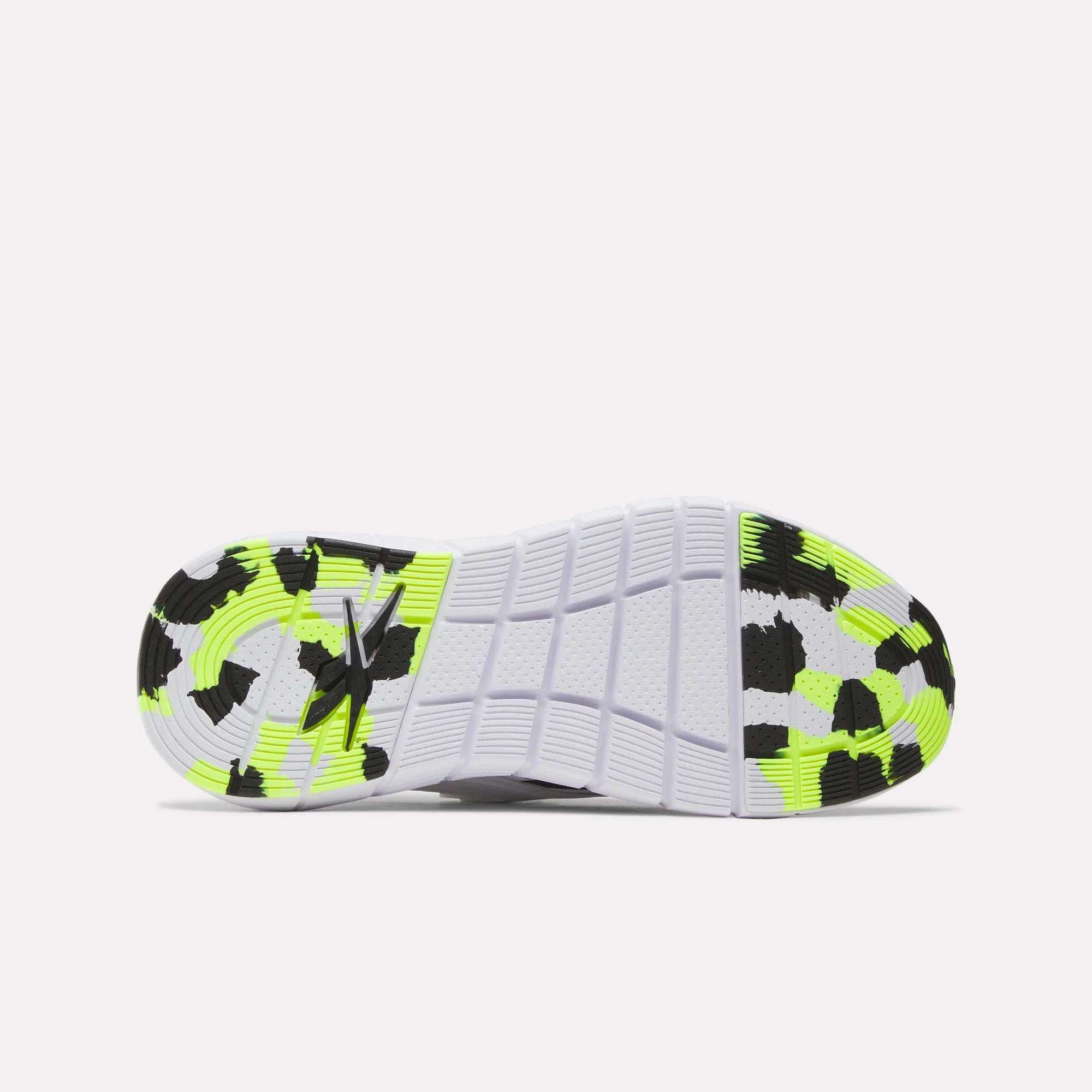 The image shows the sole of Reebok Women's Nano Gym Training Shoes, mainly white with black and neon yellow-green speckles. The tread has rectangular textures and a logo by the heel, designed for optimal grip during weight training.